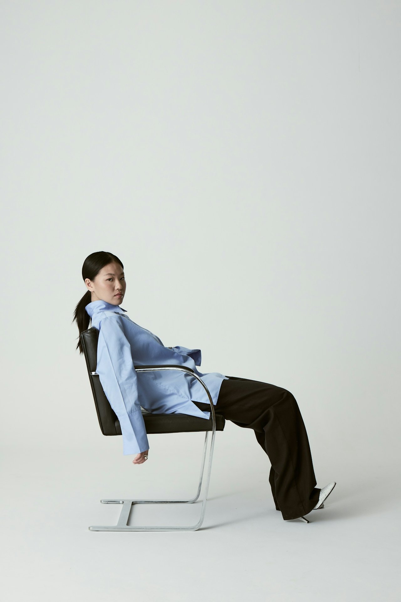 Editorial fashion photograph of a woman in a light blue blazer and wide-leg trousers seated on a sleek chrome cantilever chair, representing the premium aesthetic of Knight Theory™ creative studio.