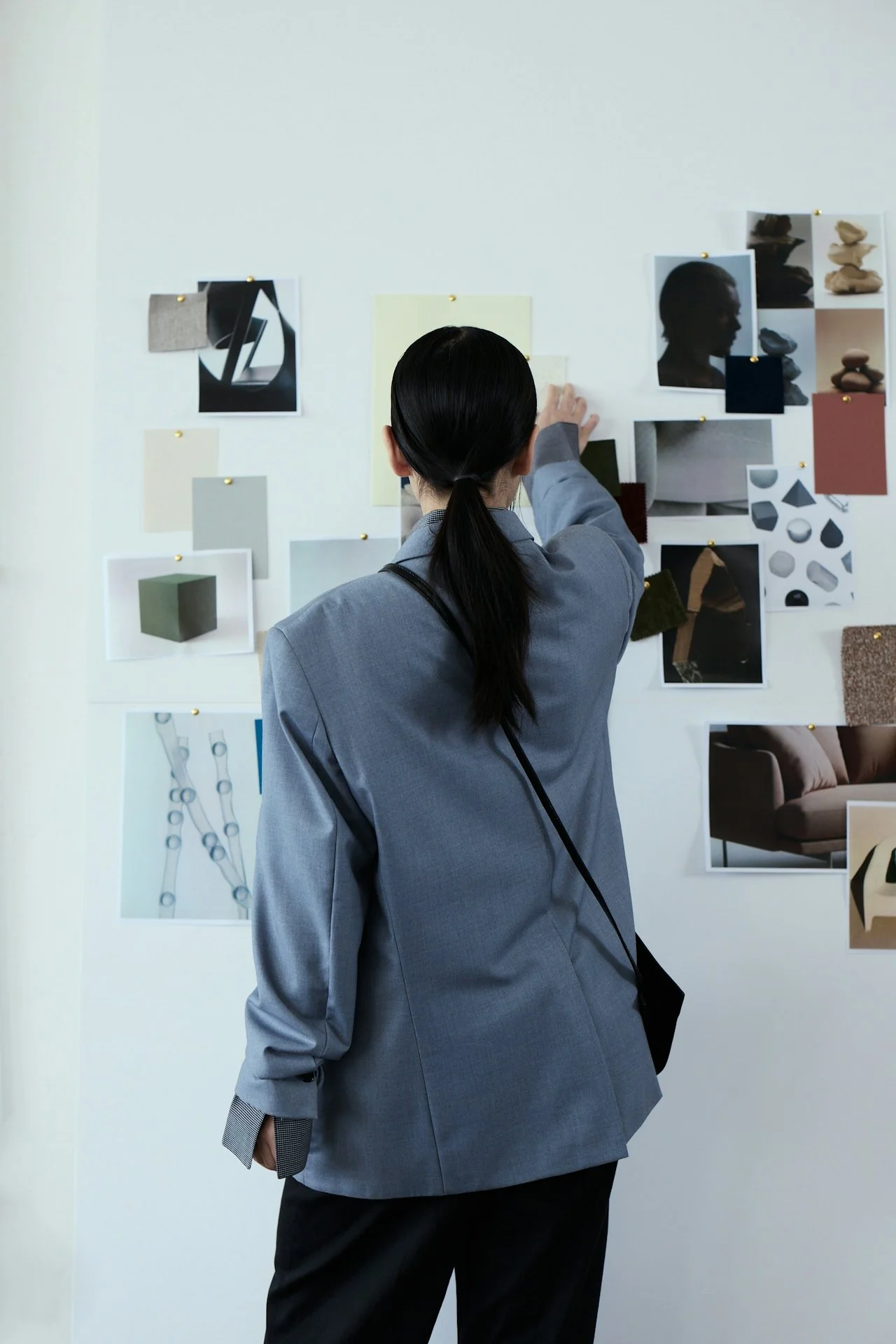 Person in a gray blazer viewing a wall of various photographs, sketches, and design images pinned to a white wall, possibly examining a creative or design project.