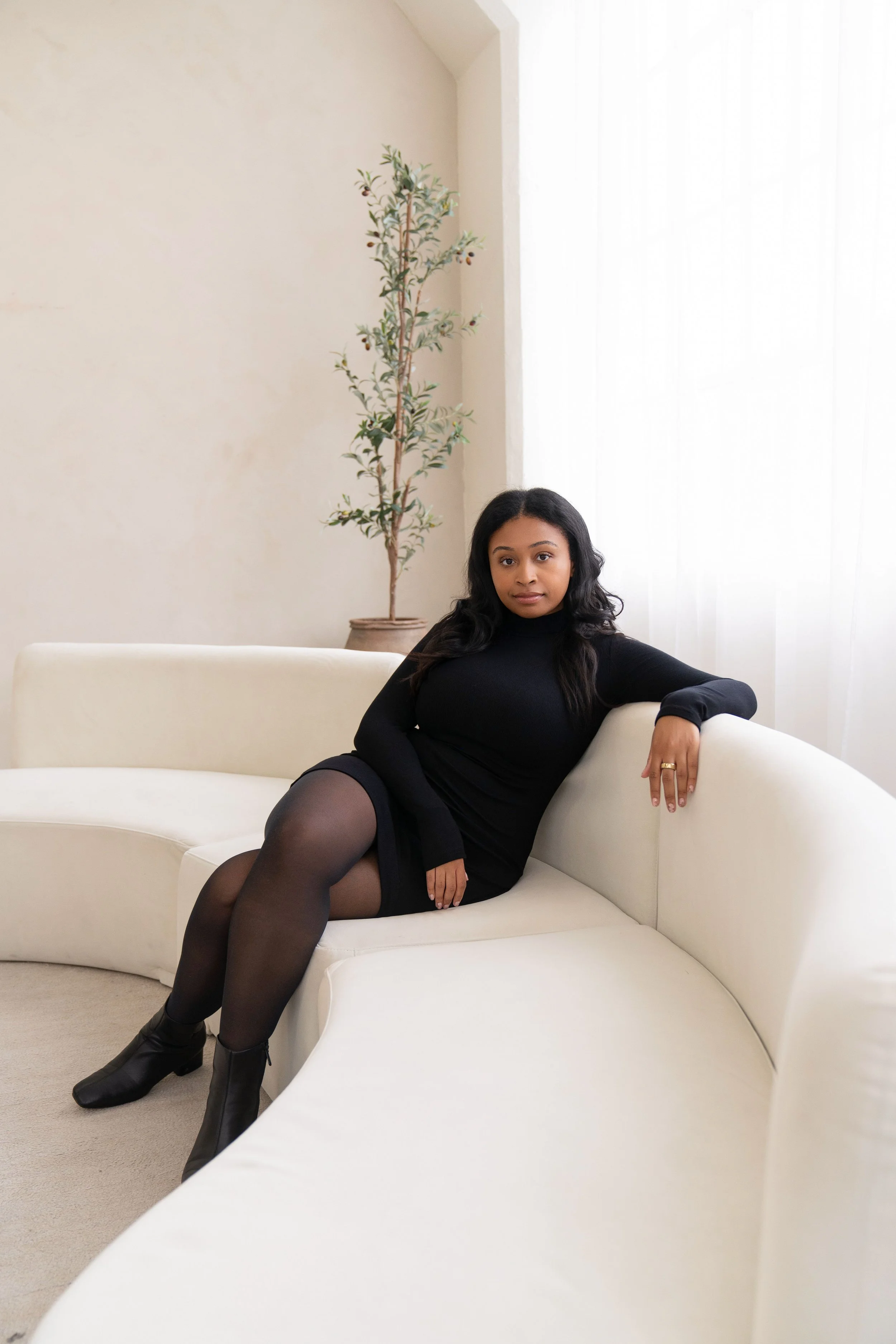 A woman in a black dress sitting on a white curved sofa in a bright room with a plant in the background.