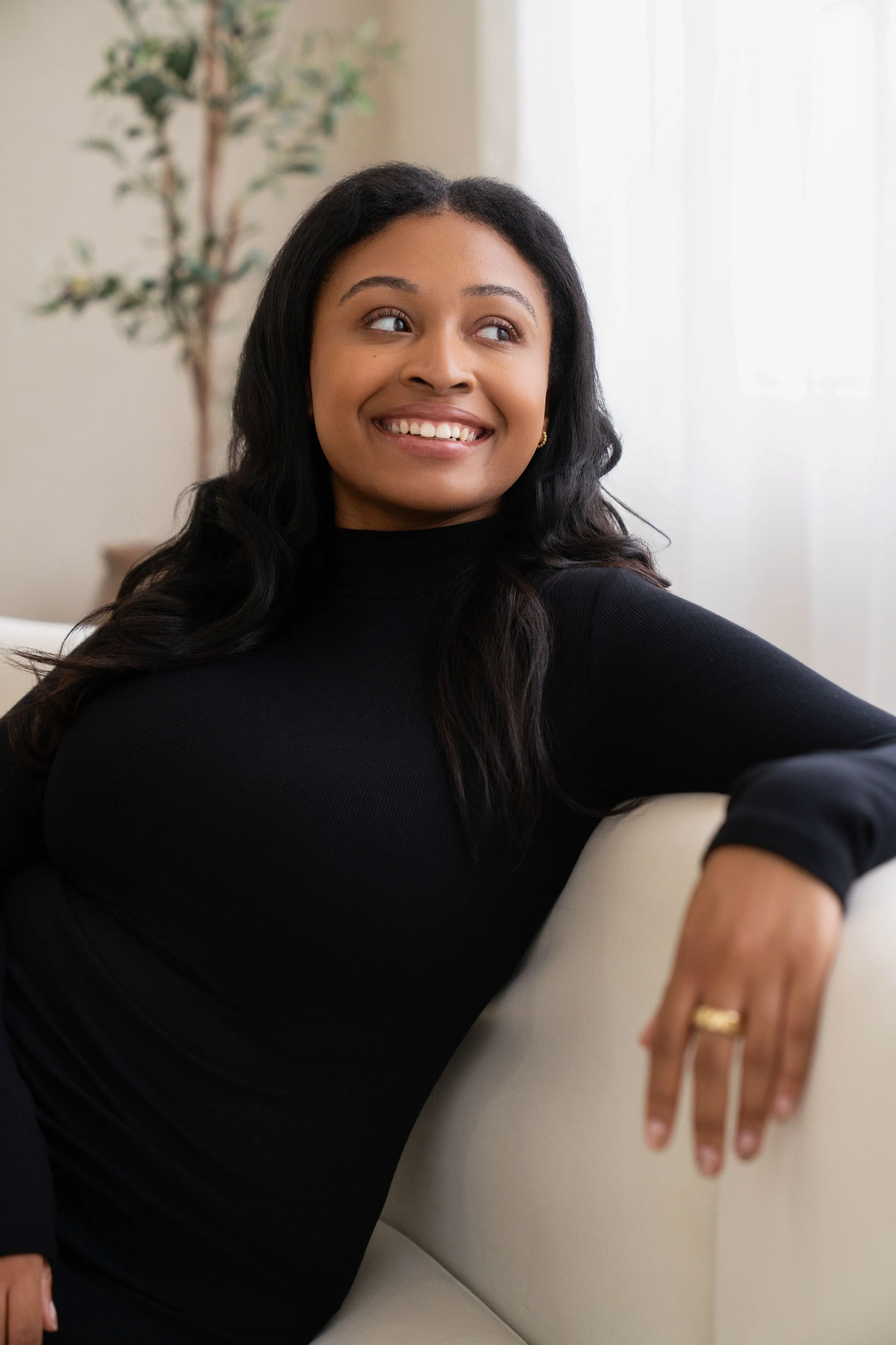A woman with long dark hair, wearing a black turtleneck sweater, smiling and sitting on a white couch with a arm resting on the armrest. There is a potted plant and a light-colored curtain in the background.