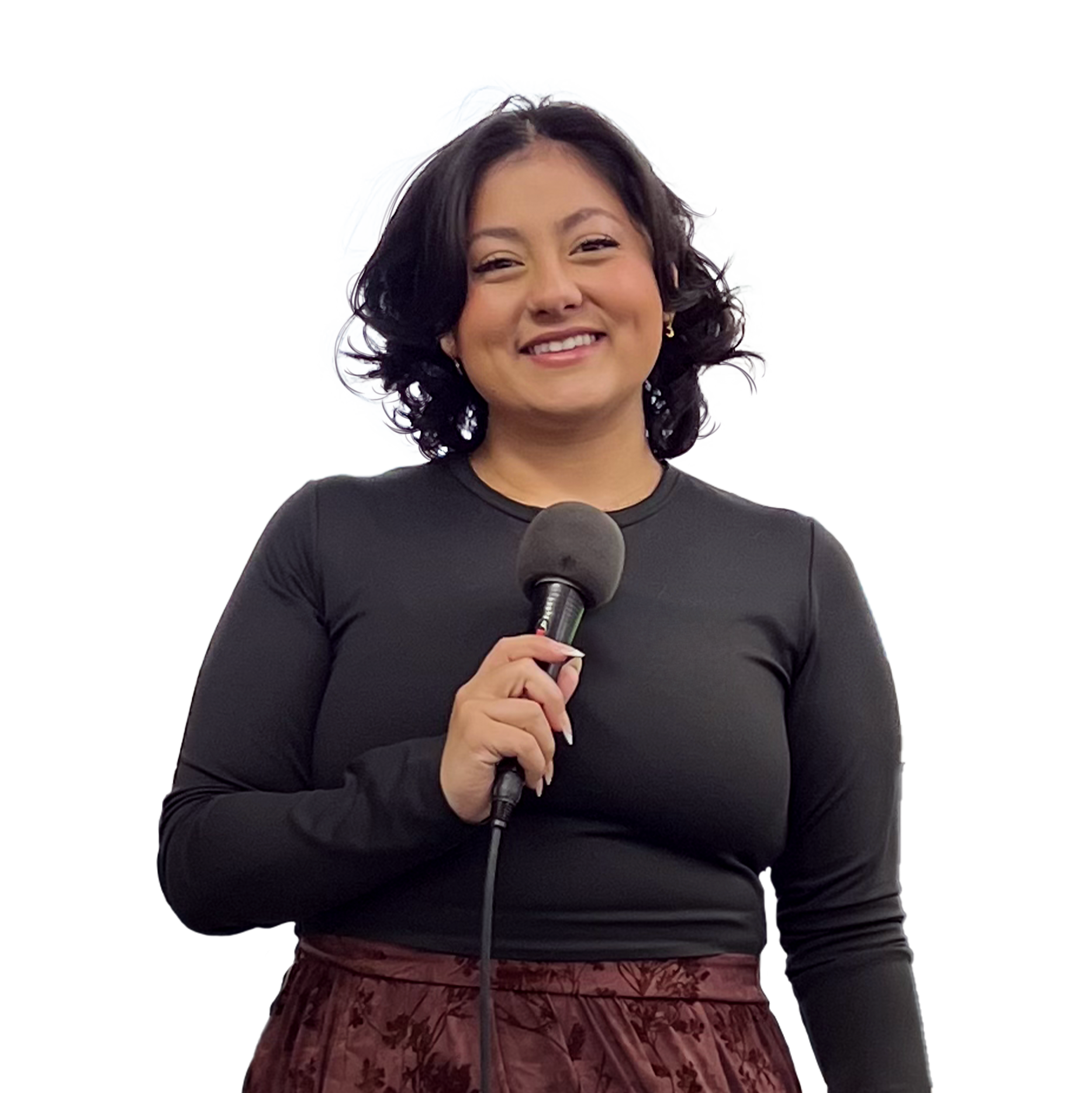 A woman with shoulder-length curly black hair, wearing a black long-sleeve shirt and a red patterned skirt, smiling and holding a microphone.