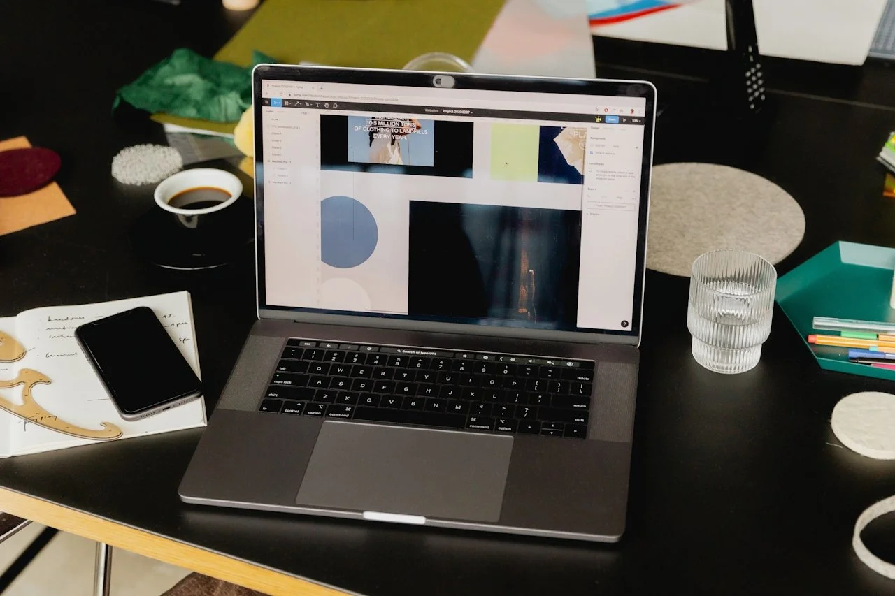 Laptop displaying a graphic design program with colorful brand assets on a dark desk alongside a coffee and phone, representing the custom Squarespace website design and development phase at Knight Theory™ Los Angeles.