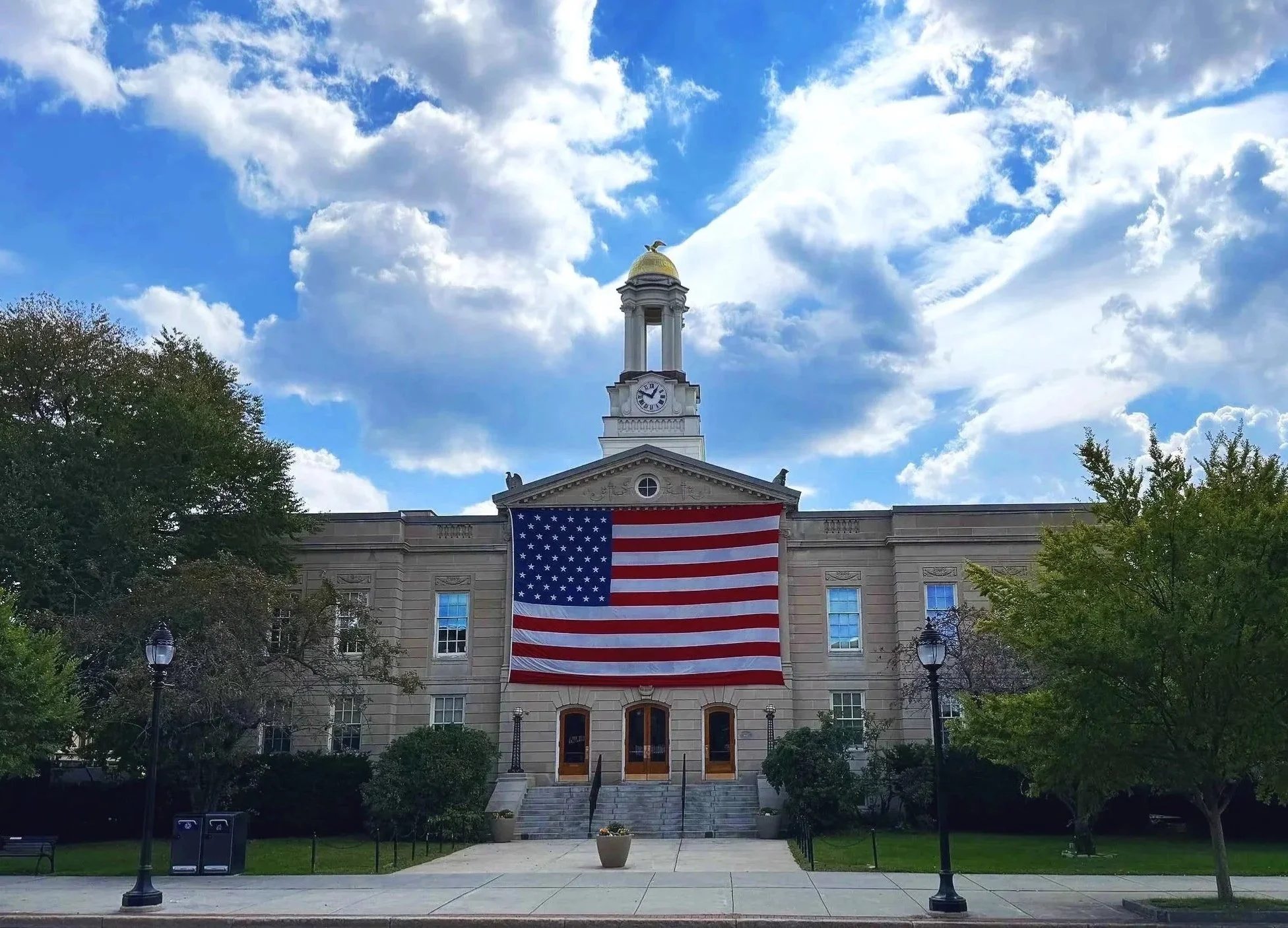 A large American Flag draped over the entrance to Waltham City Hall with lush green trees in front and sun-kissed clouds above in a blue sky