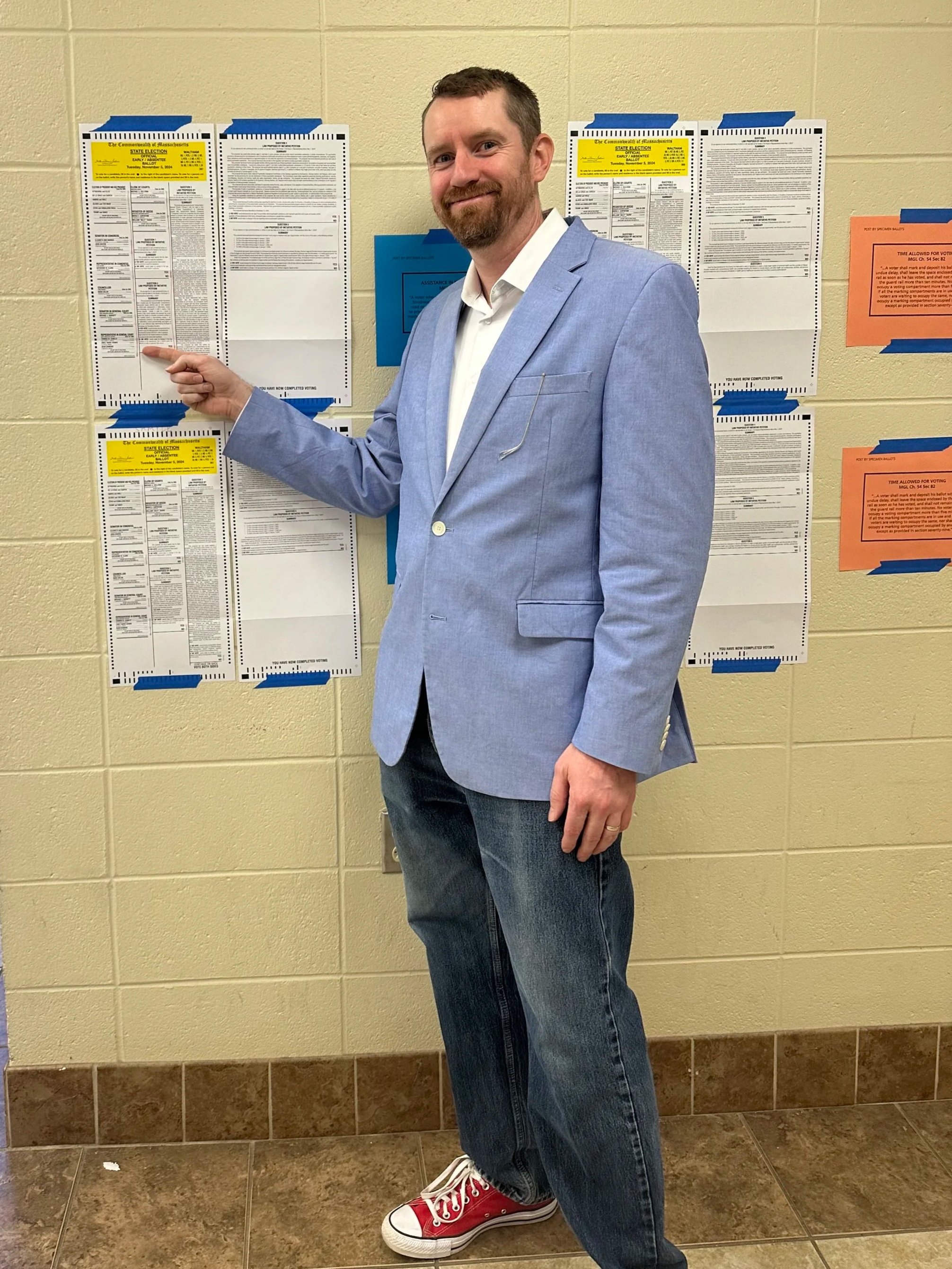 Sean Diamond wearing a light blue blazer, white button down shirt, blue jeans, and red converse sneakers. Sean is pointing to his name on the 2024 sample ballot when he ran for Waltham State Rep as an independent candidate.