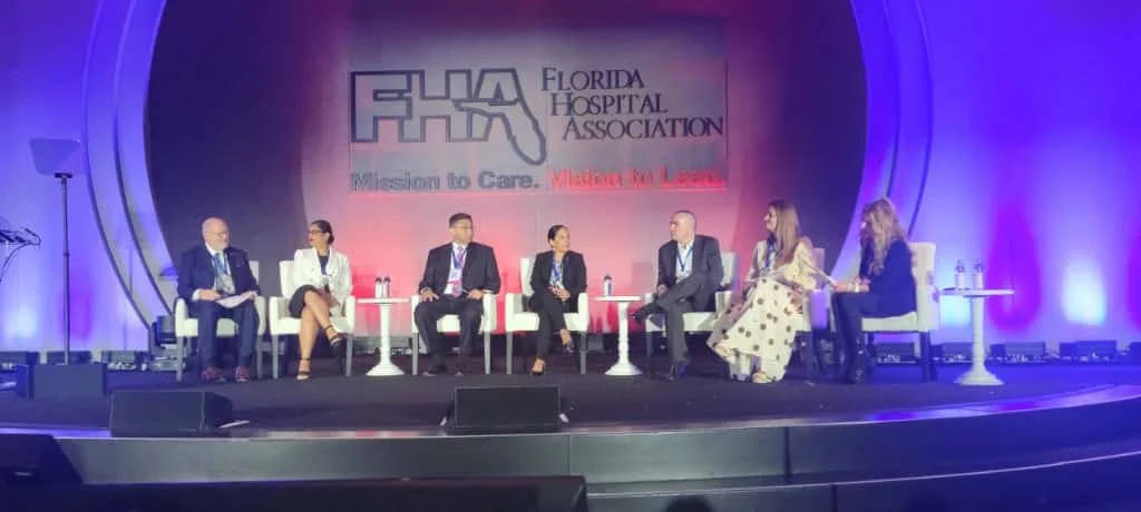 Panel of seven people seated on a stage with a Florida Hospital Association banner in the background, participating in a conference or discussion.