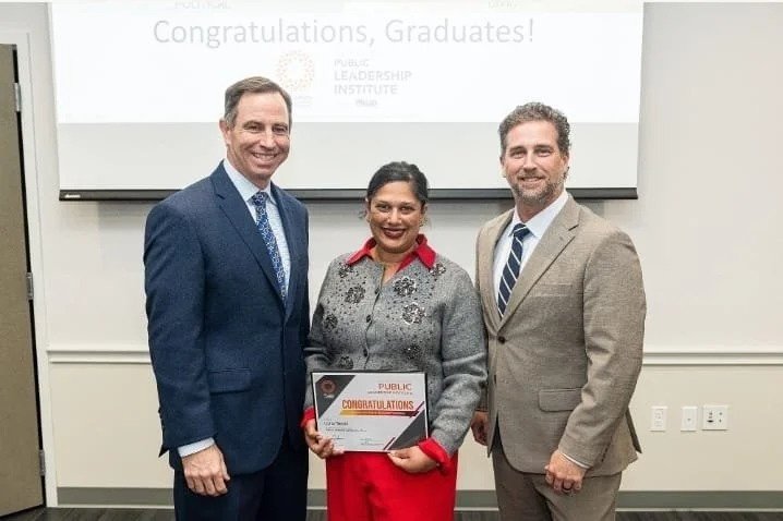 Three people standing together in a room, smiling at the camera. The woman in the middle is holding a certificate. Behind them is a screen with a message that says, "Congratulations, Graduates!"