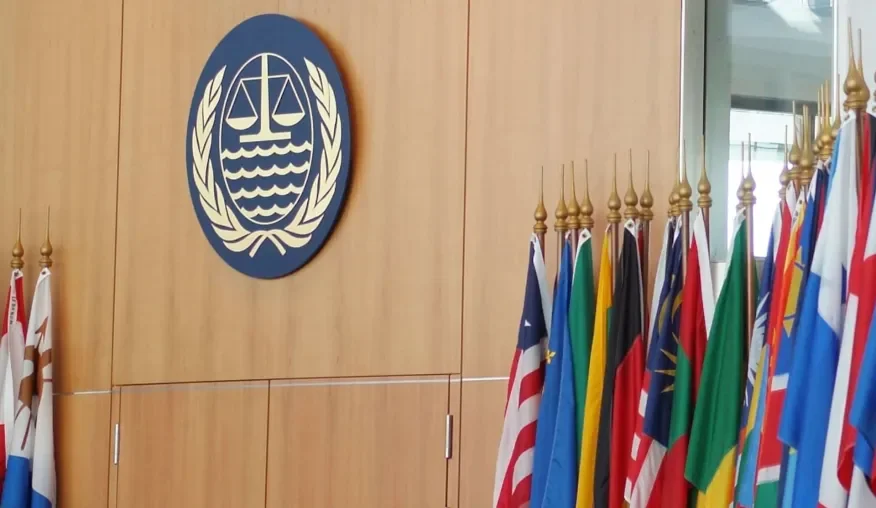 A wall with a large emblem featuring scales of justice, waves, and laurel branches, and a row of international flags on gold poles.