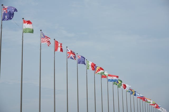 Multiple international flags flying on flagpoles against a cloudy sky.