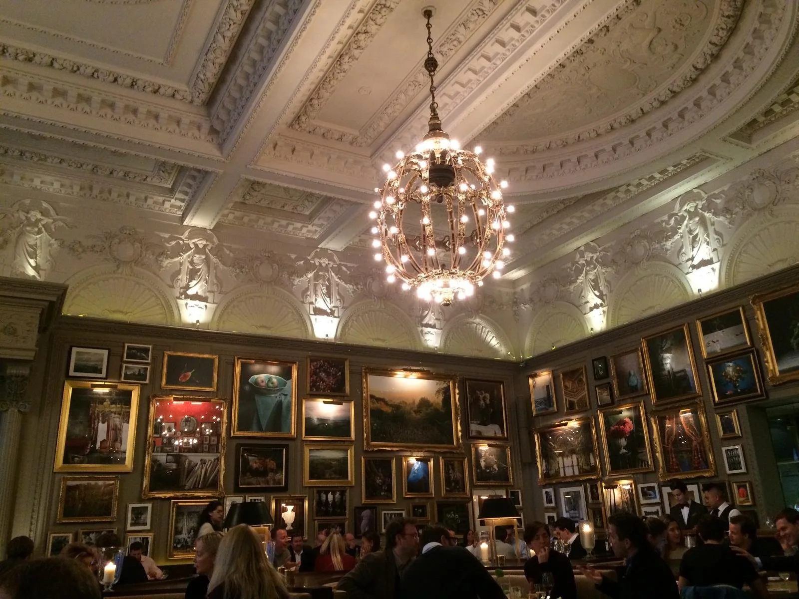 An elegant dining room with a ornate ceiling, a crystal chandelier, and walls lined with framed paintings. People are sitting at tables enjoying a meal.