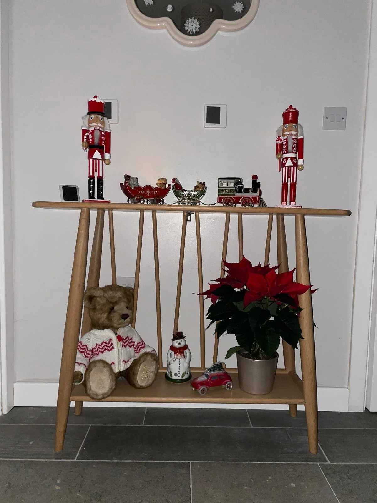 Decorative Christmas display on a wooden table with nutcracker figurines, a toy train, teddy bear, poinsettia plant, snowman figurine, and a small toy car against a white wall.