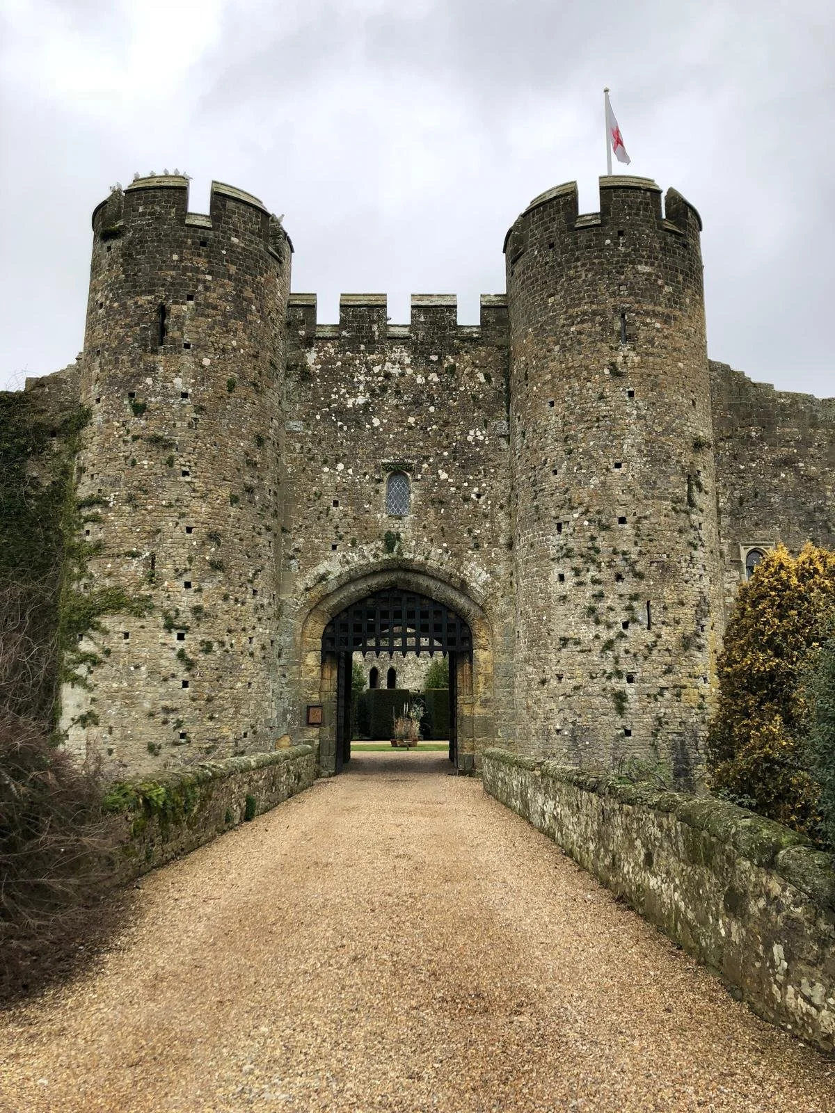 Ancient stone castle with a gateway and two cylindrical towers, one with a flagpole flying a white flag with a red cross, overcast sky.