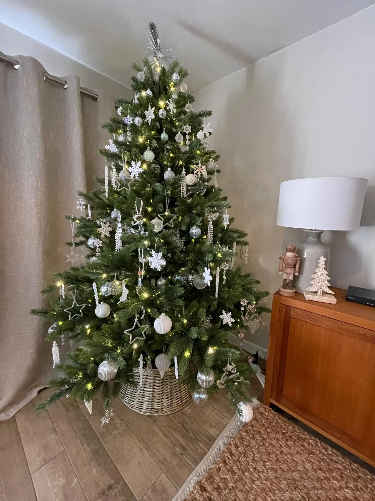 A decorated Christmas tree with white and silver ornaments next to a wooden side table with a white lamp and holiday decorations.