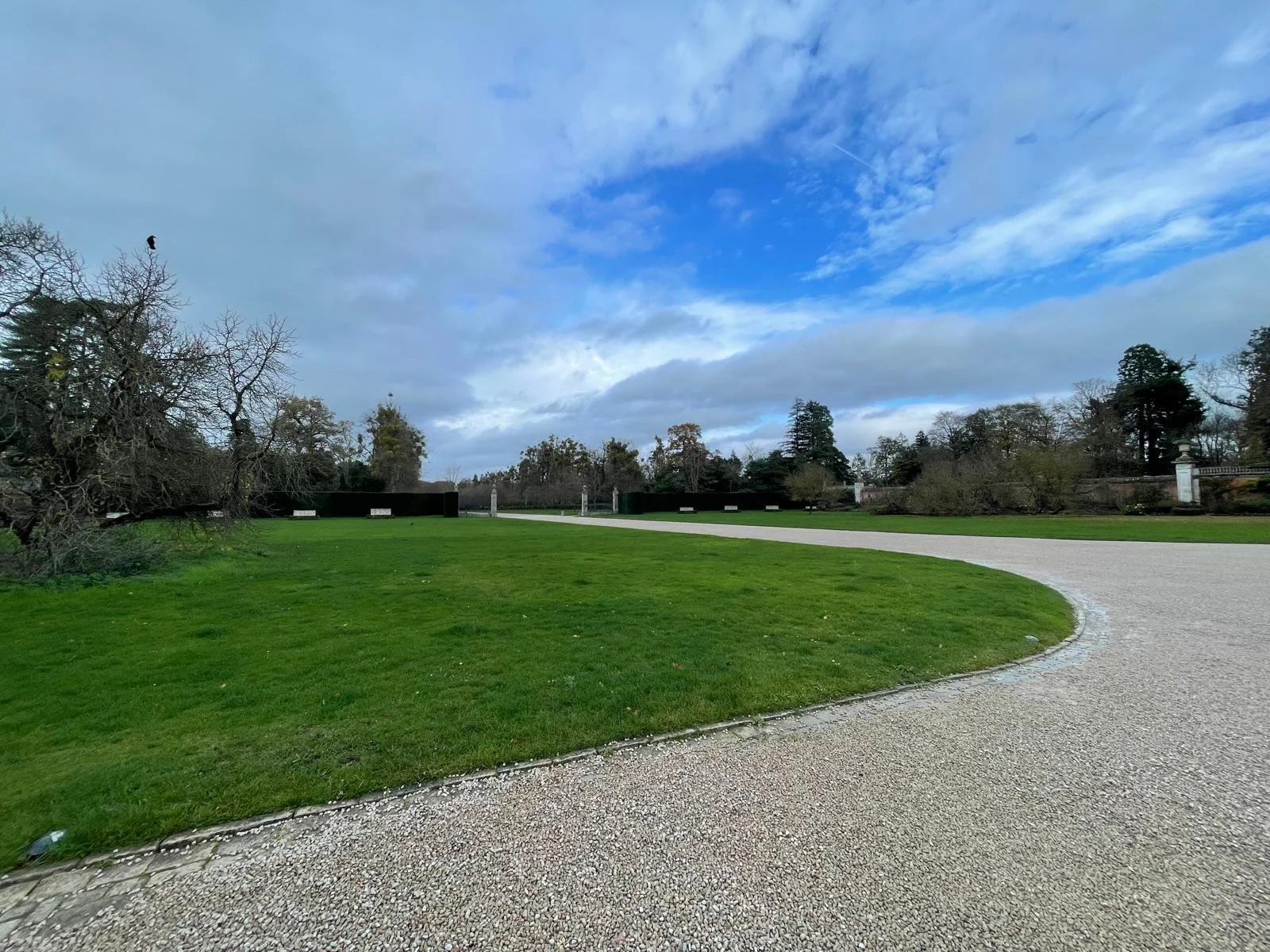 A park with green grass, a curved gravel pathway, leafless trees, and a partly cloudy sky.
