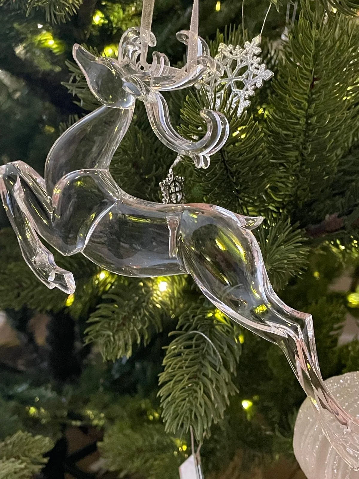 A transparent glass reindeer ornament hanging on a Christmas tree with green branches and warm yellow lights.