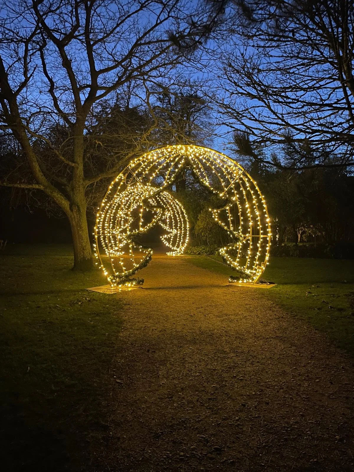 Decorative holiday light installation of two large spheres with stars inside, illuminated with yellow lights, located outdoors on a pathway at dusk or night with trees in the background.