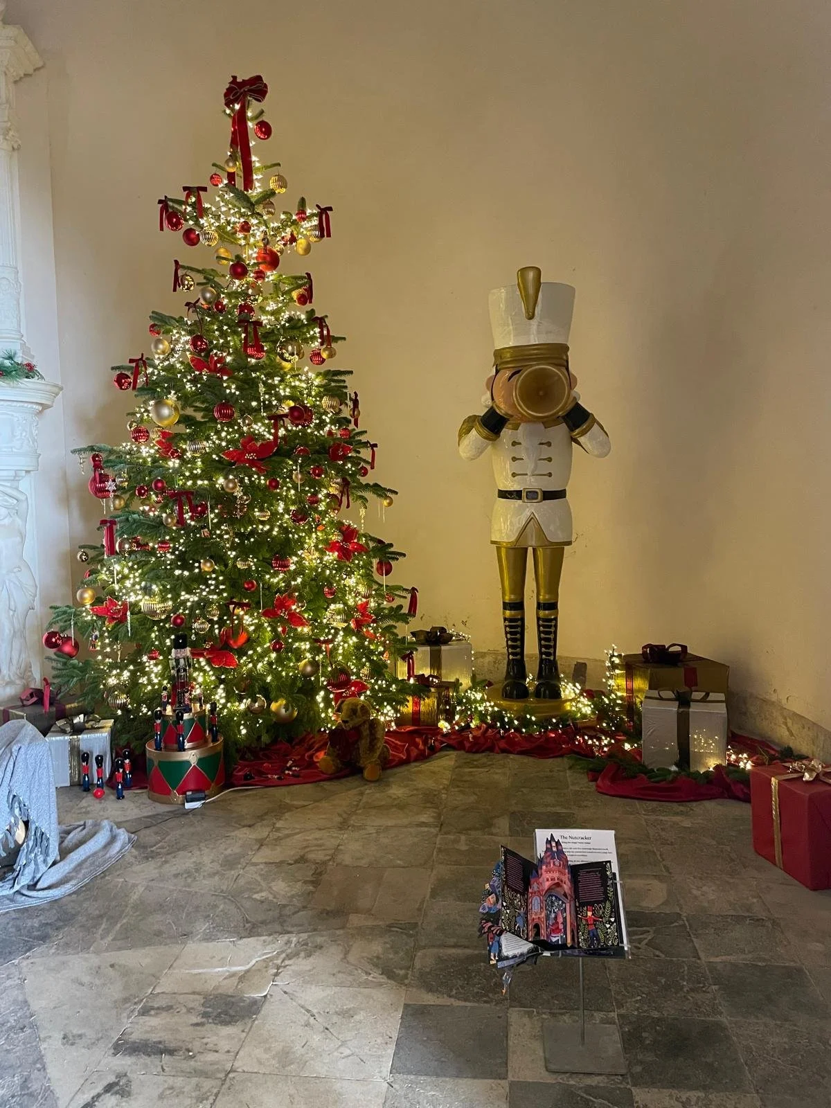 A decorated Christmas tree with red, gold and silver ornaments and white lights stands next to a large Nutcracker figure. Wrapped presents are placed under the tree, and a few toys and gifts are arranged around the base. A small, open holiday card di