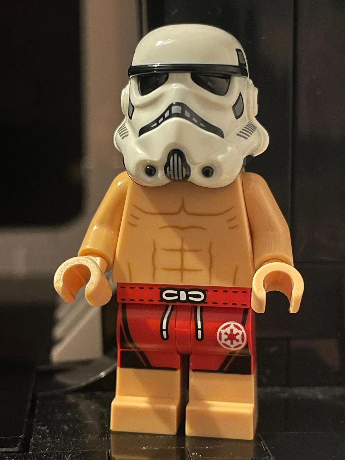 A LEGO minifigure with a Stormtrooper helmet and a muscular torso wearing red and black shorts with a white Imperial logo.