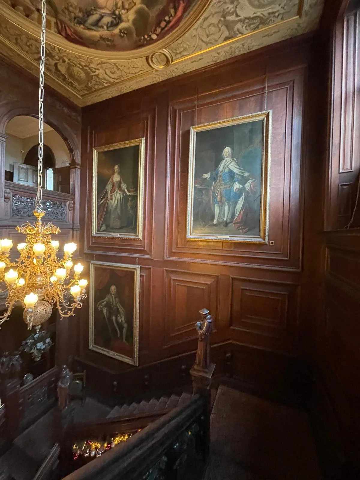 A grand staircase hall with wooden paneled walls decorated with large framed portraits of historical figures. A large chandelier with multiple lamps hangs from the ceiling, which features intricate painted artwork and gold accents.