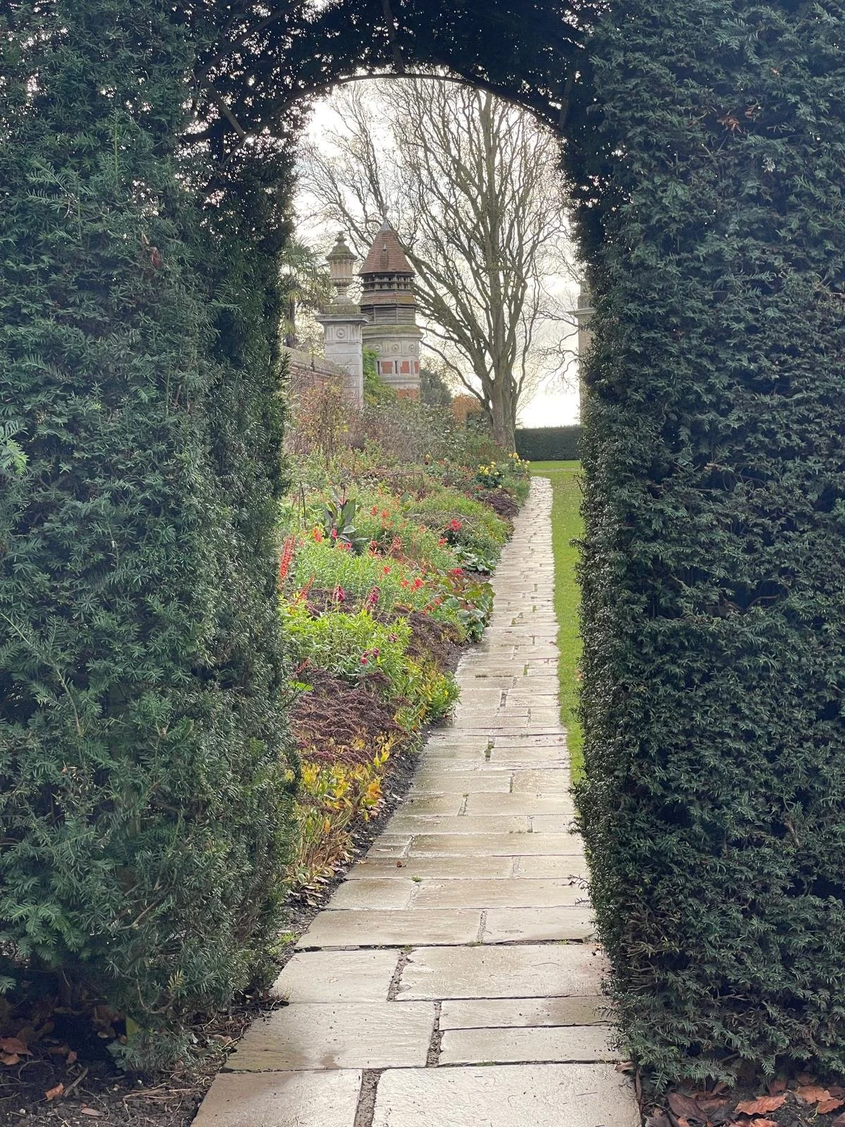 A stone-paved garden pathway viewed through an archway made of tall, dense green hedges, with a flower bed on the left and a large tree in the background.