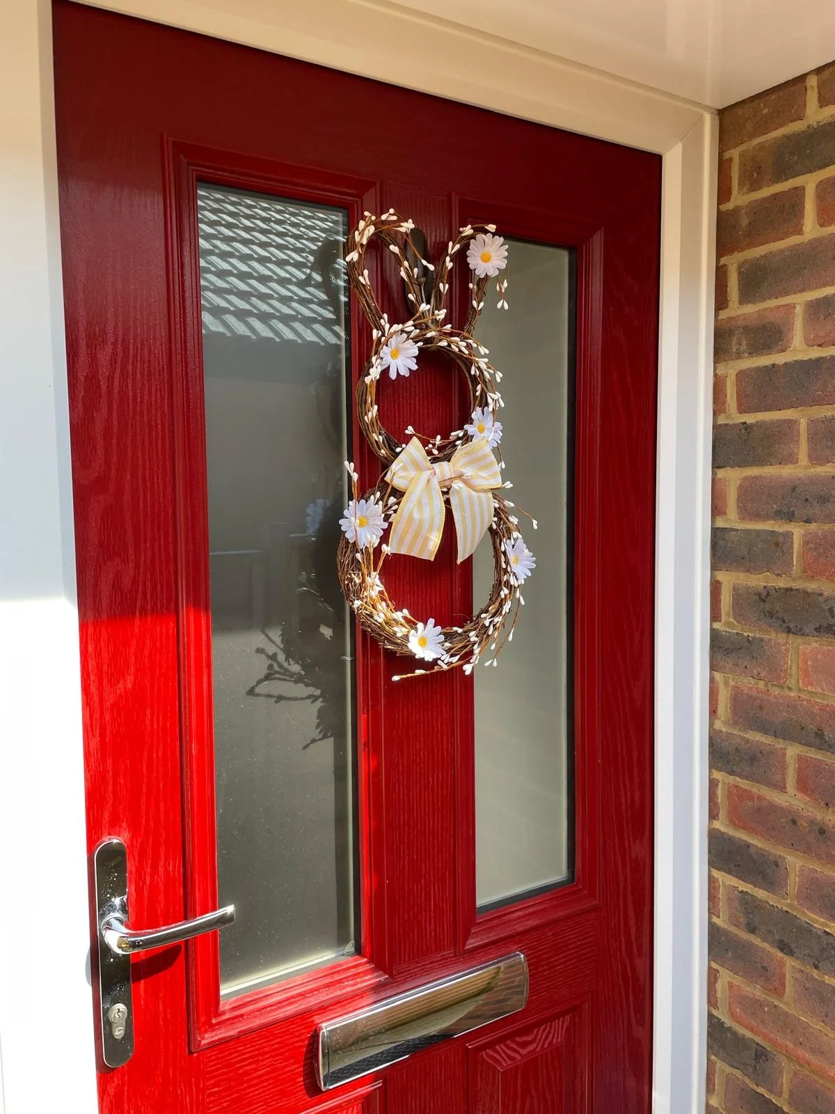 Red front door decorated with a wreath featuring two intertwined circles, daisies, a yellow and white striped bow, and small white lights.