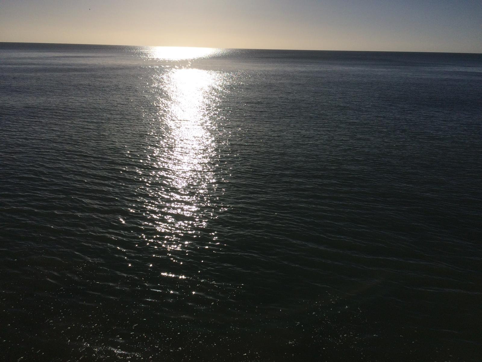 The sun reflects on the ocean's surface, creating a bright, shimmering trail of light extending from the horizon to the foreground, with calm waves under a clear sky.