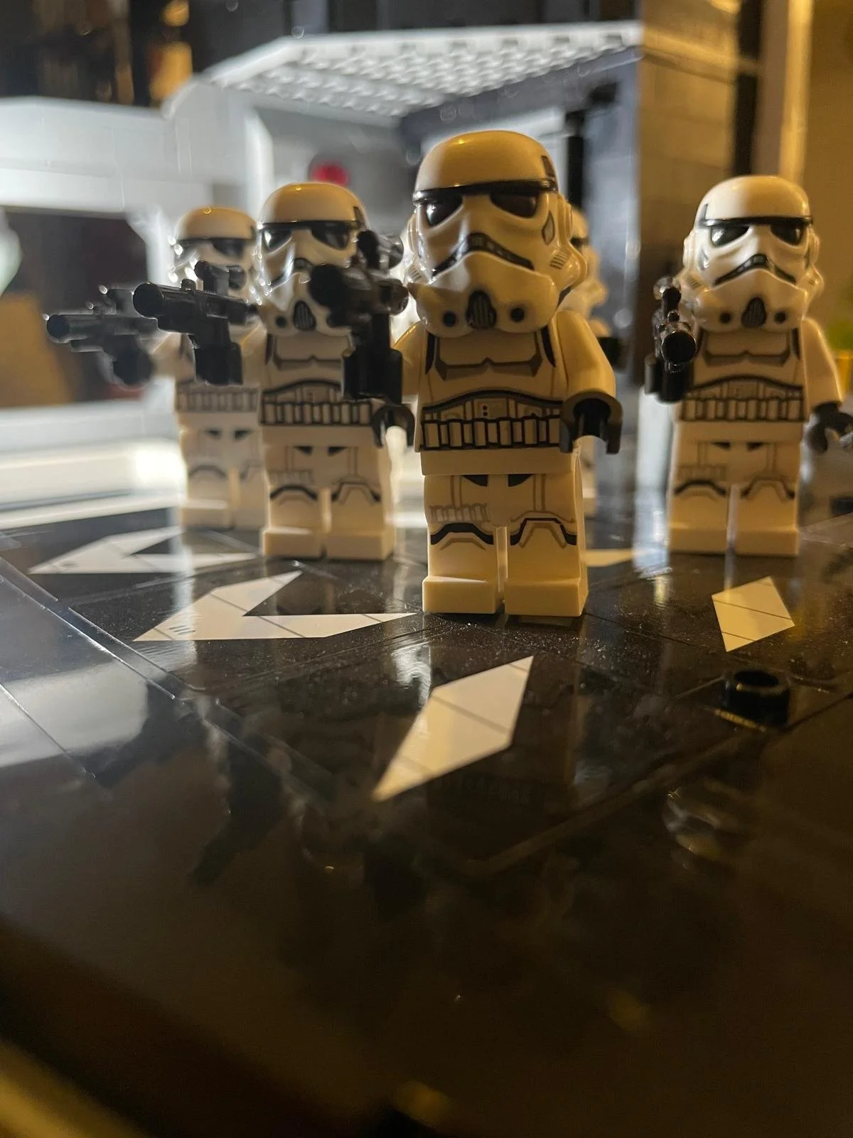 A group of LEGO Star Wars stormtrooper figurines standing on a table with a black and white geometric pattern, with some figures holding small black blaster rifles, and a blurred background of a room.