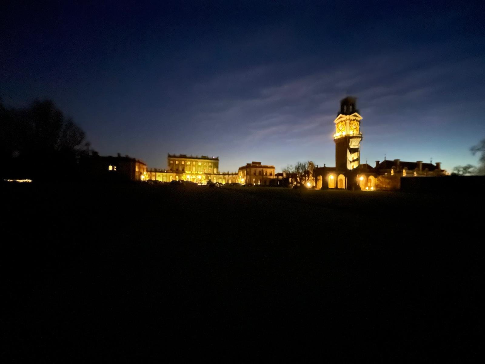 Nighttime view of a historic building with a tall clock tower, illuminated with warm yellow lights, against a dark sky.