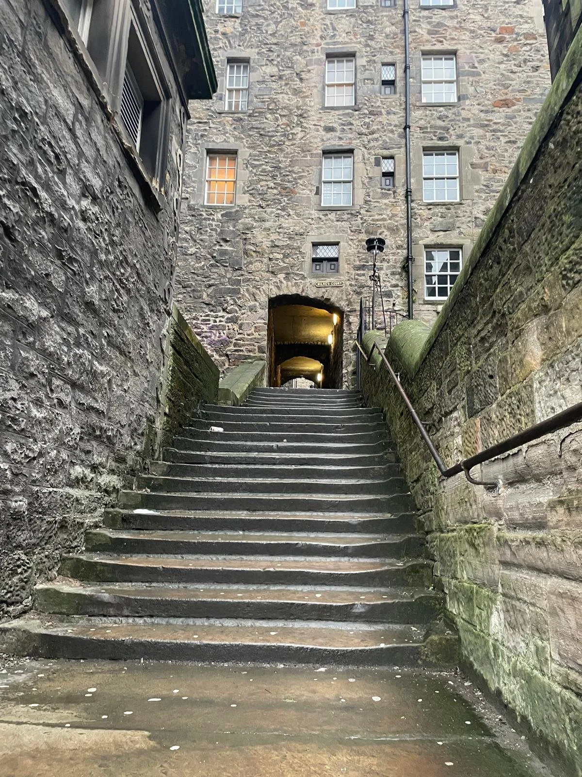 A stone staircase leading up to an archway and an old stone building with multiple windows, some lit from inside, in an alleyway or courtyard.