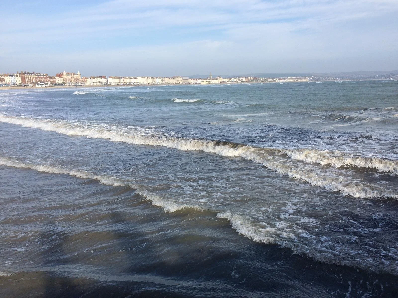 View of a beach with gentle waves and a distant shoreline with buildings under a partly cloudy sky.