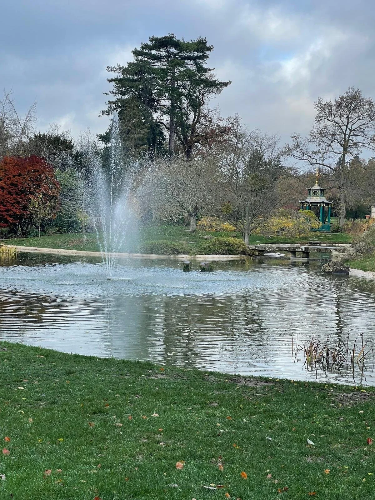 A peaceful park scene with a pond and a fountain spraying water, surrounded by trees in fall foliage, a small bridge, and a decorative gazebo. Overcast sky.