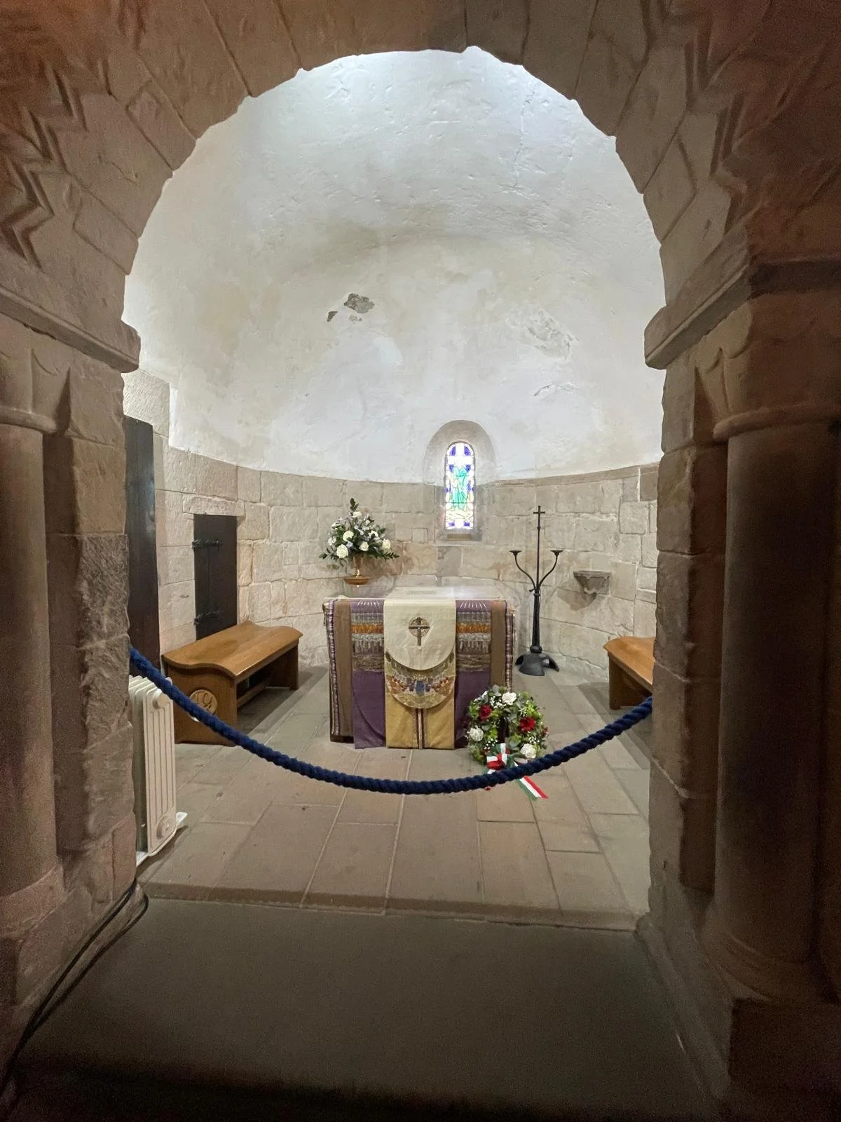 Small chapel with altar, stained glass window, flowers, cross, and candelabra, viewed through an arched stone doorway.