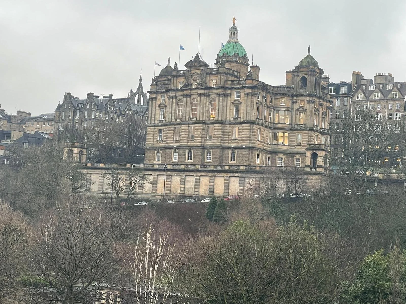Historical stone building with domed towers and flags, surrounded by leafless trees and other buildings, under a cloudy sky.