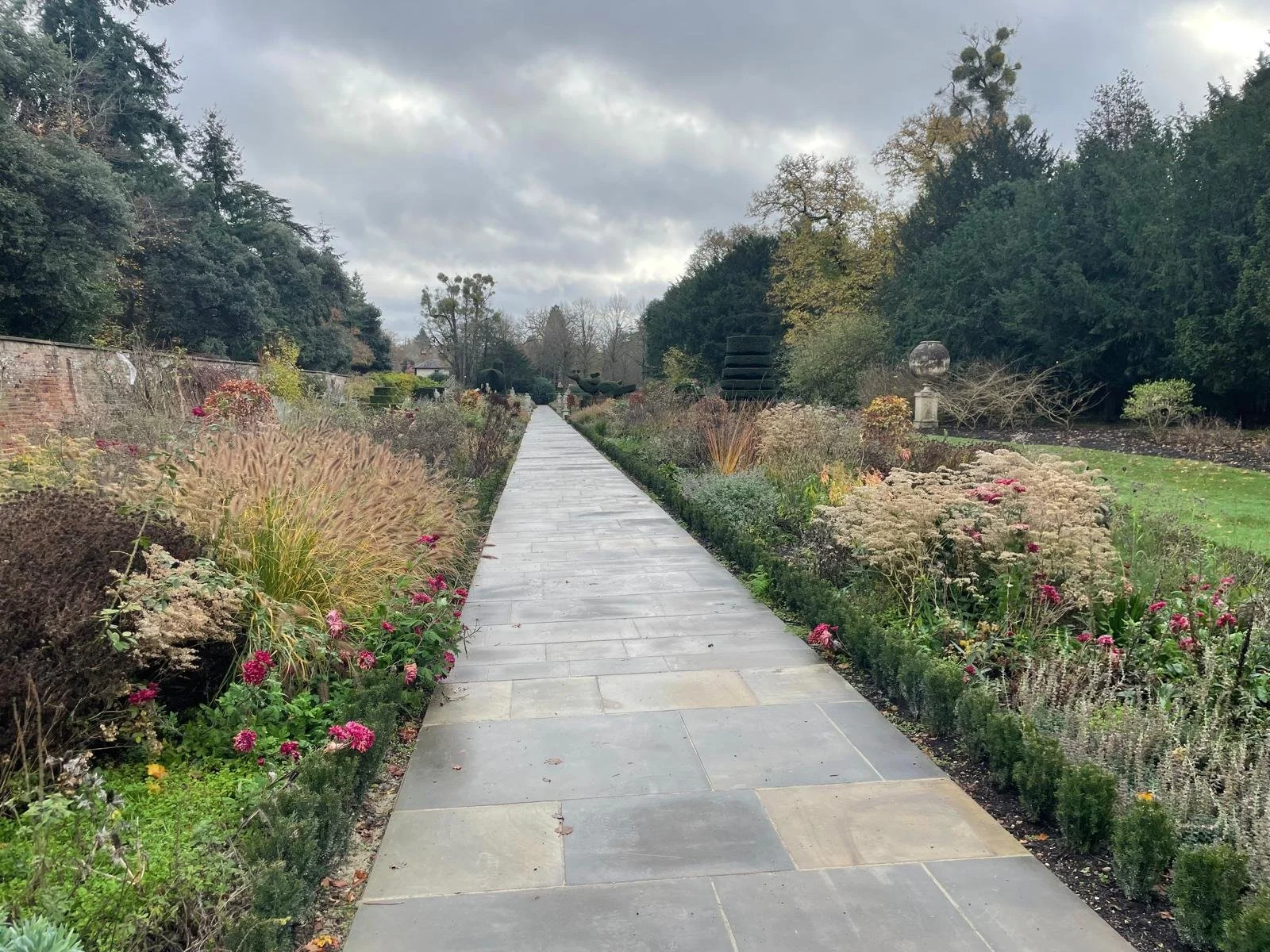 A stone pathway running through a landscaped garden with various flowers and shrubs on either side, bordered by trees and a brick wall on the left, under a cloudy sky.