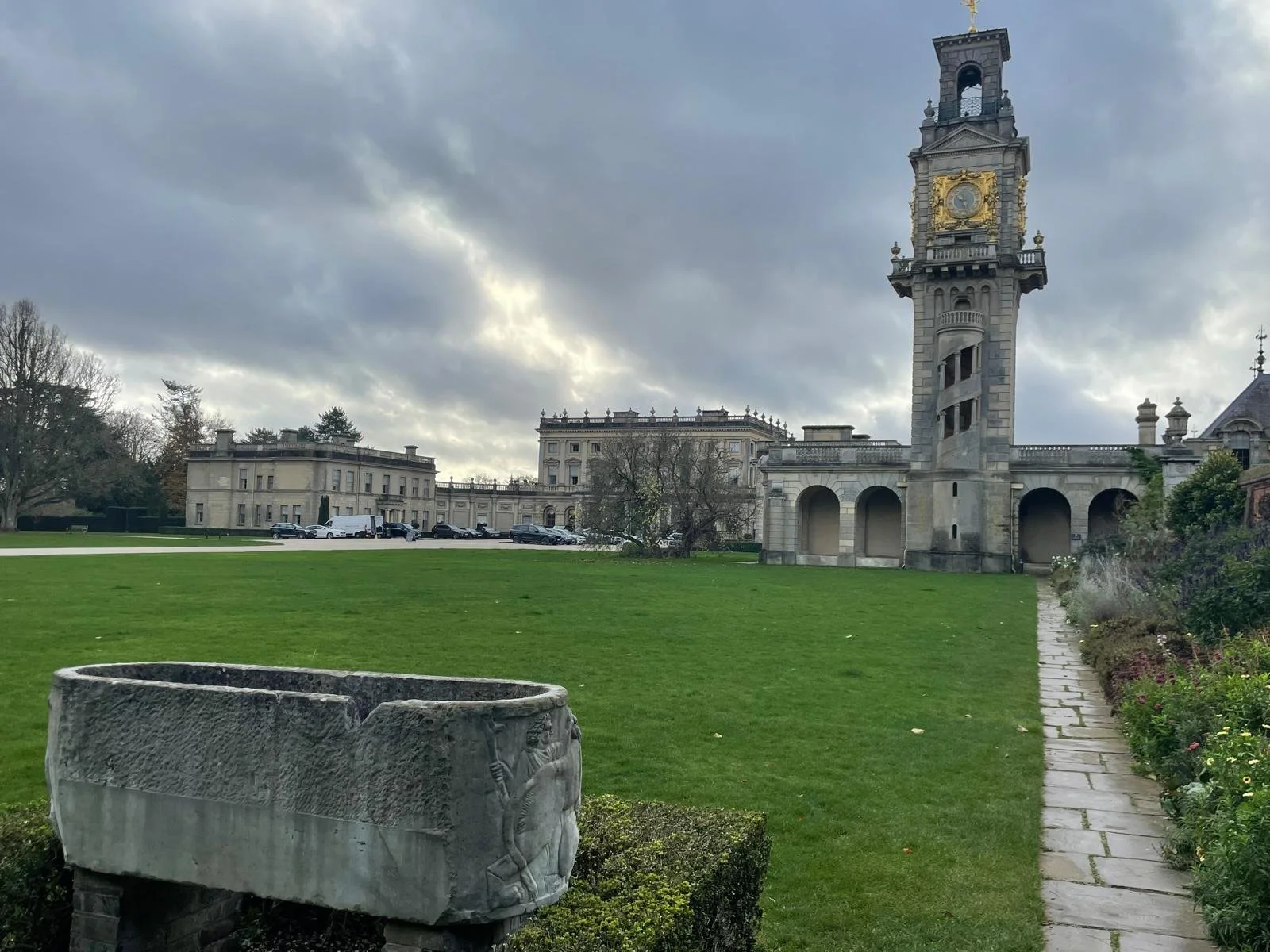 A historic building with a clock tower on a cloudy day, surrounded by a well-manicured lawn and a stone pathway, with parked cars and trees in the background.