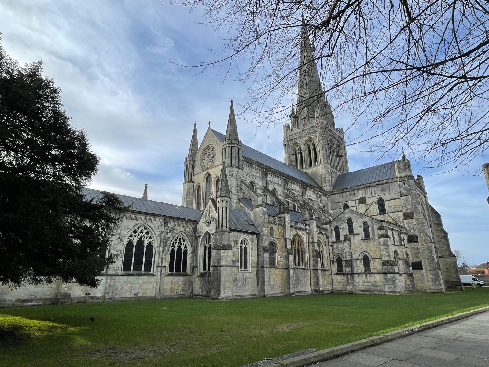 A large Gothic-style stone church with tall steeples, arched windows, and a clock on the facade, set against a partly cloudy sky with leafless tree branches in the foreground.