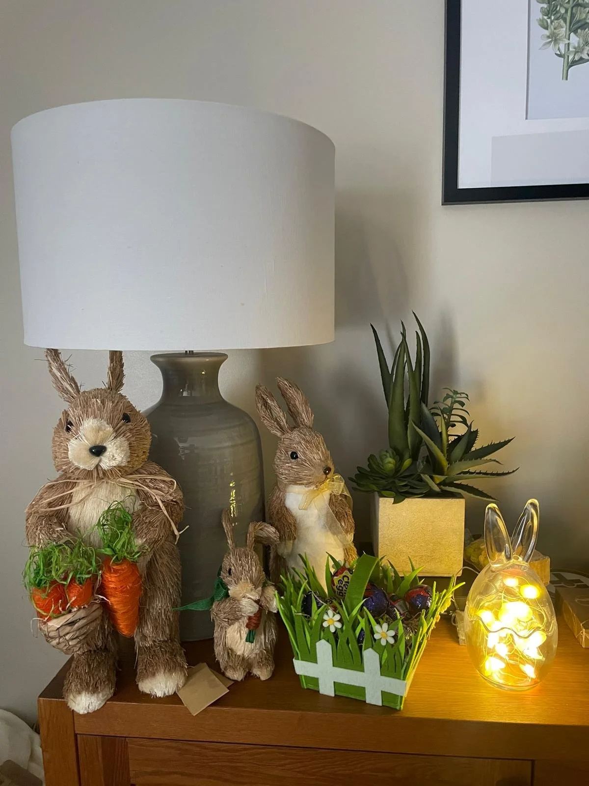 Decorative Easter-themed display on a wooden table, featuring a white lampshade with bunny figurines made of straw in front, green plants including succulents in yellow and white pots, a glass bunny-shaped lantern with lights, and a framed botanical 