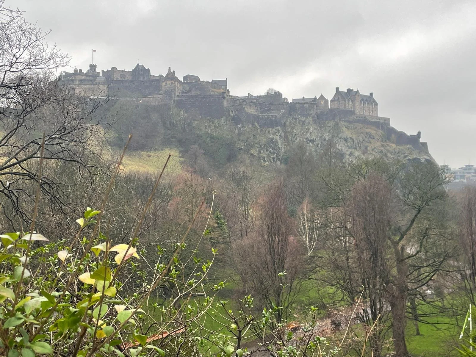 A castle situated on a hilltop overlooking a wooded area with leafless trees and green patches, under a cloudy sky.