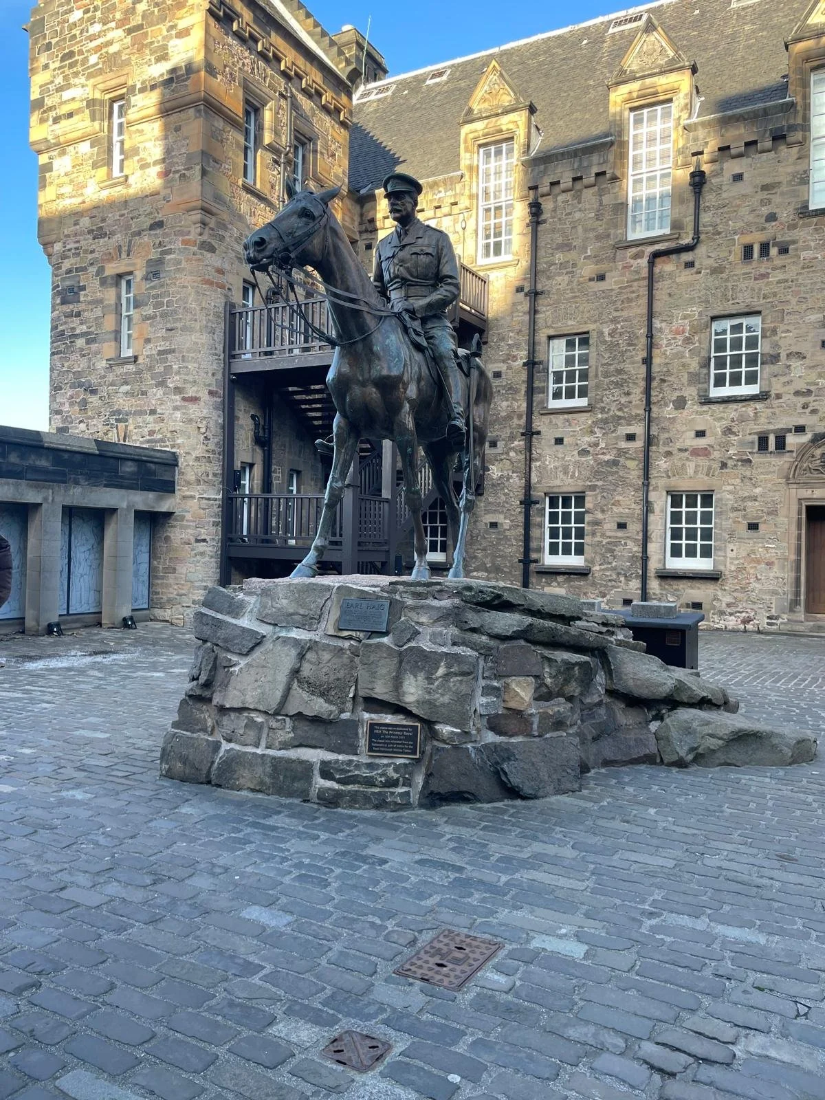 A bronze statue of a man on horseback, elevated on a stone platform, located in a cobblestone courtyard with historic stone buildings in the background.
