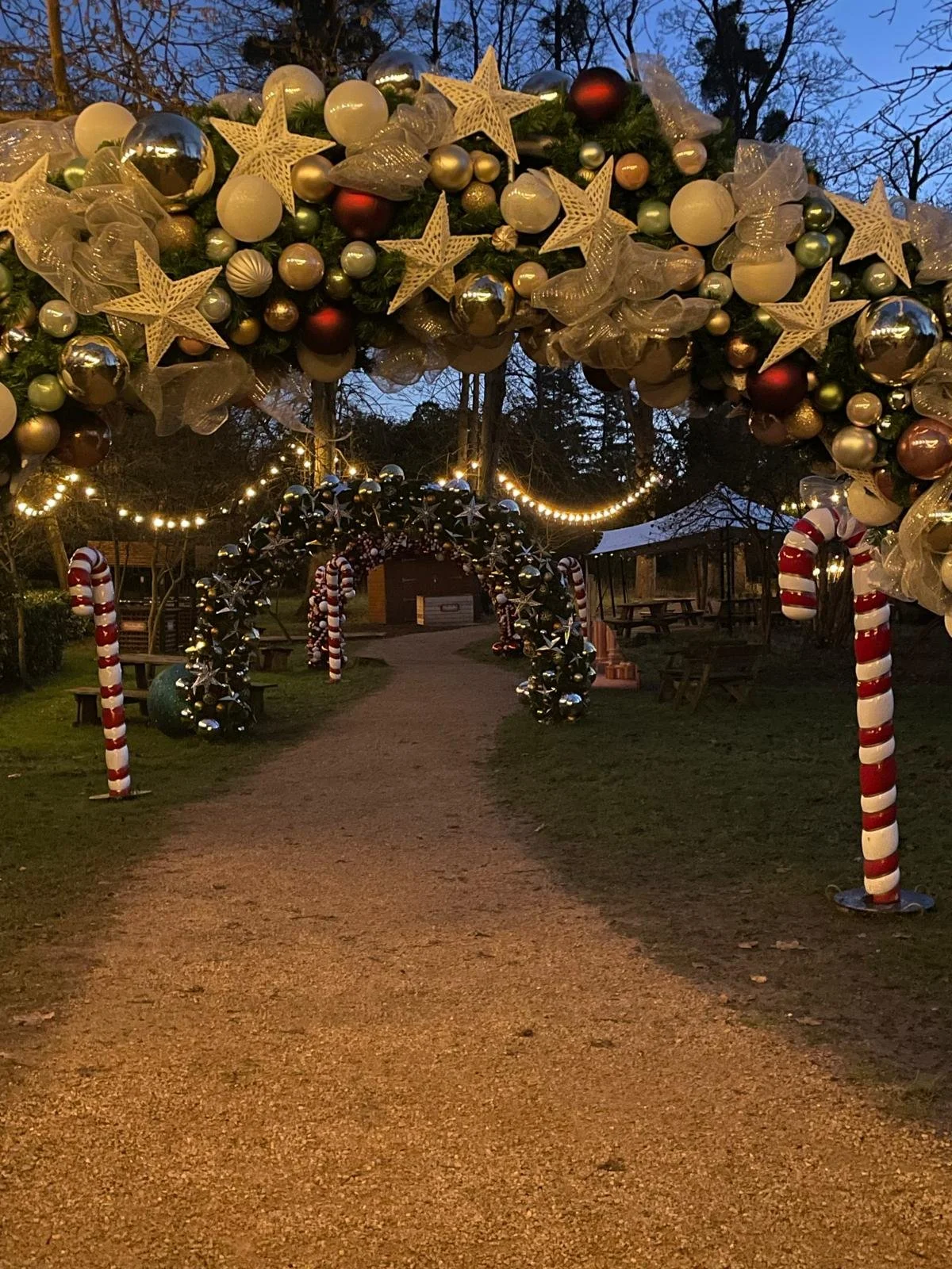 Outdoor Christmas decoration with arched gateways adorned with baubles, stars, and ribbons, along with peppermint-striped poles and candy cane decorations, illuminated during evening.