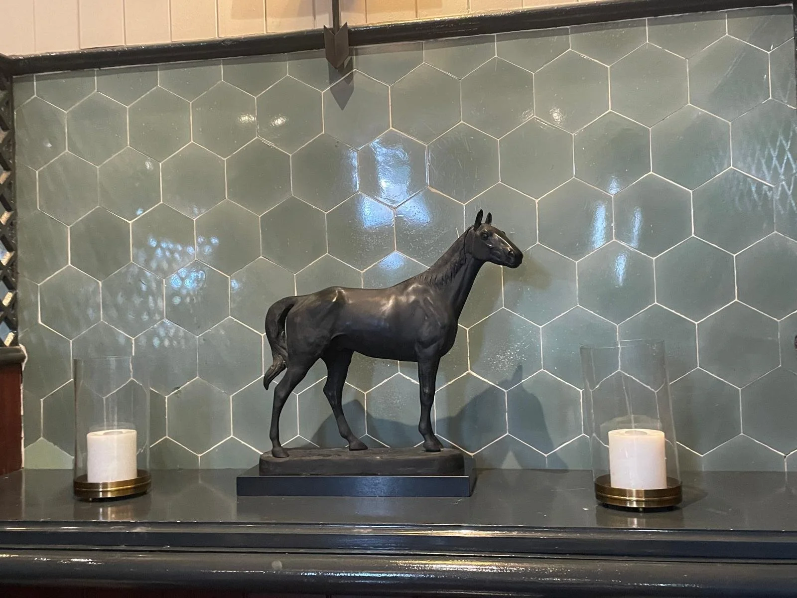 A black horse sculpture on a black base flanked by two white candles with gold bases, against a green hexagonal tile wall.