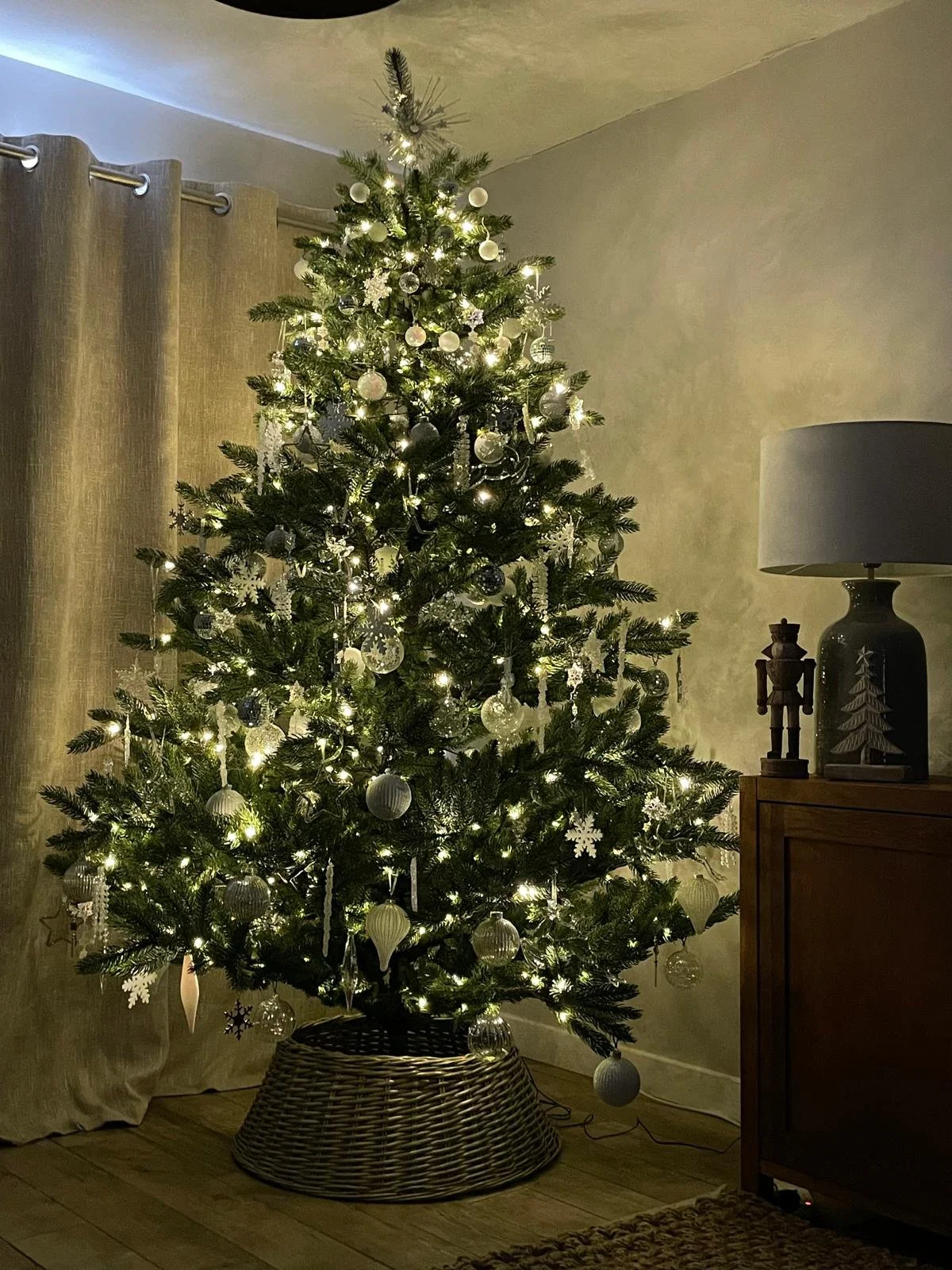 Decorated Christmas tree with white and silver ornaments, lights, and a topper, standing in a living room near a window with beige curtains and a side table with decor, including a lamp and ornaments.