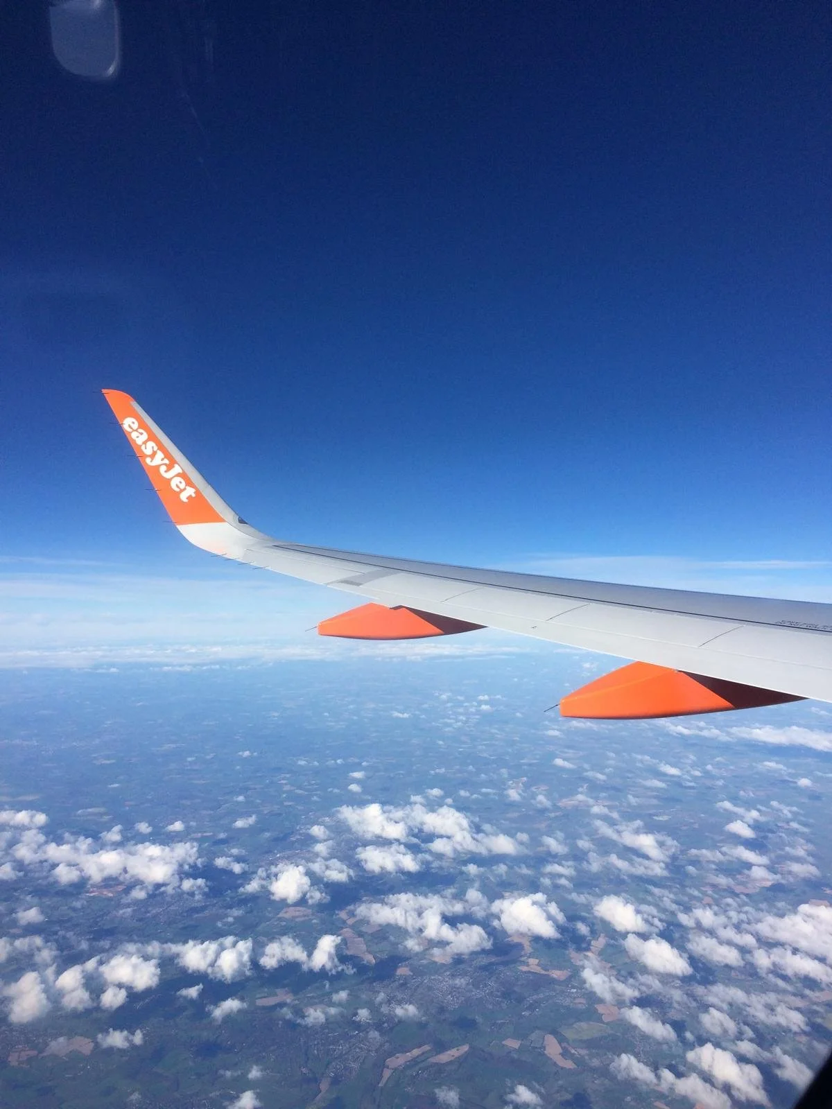 View from an airplane window showing the wing labeled 'easyJet' flying above clouds and land with a clear blue sky.