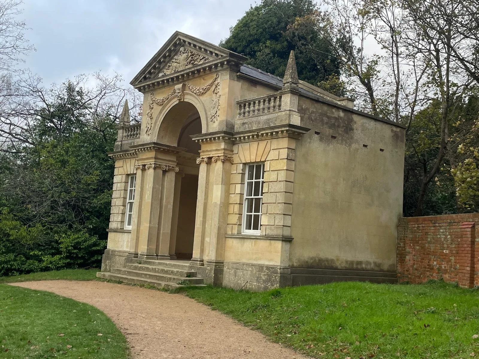 An historic, small, classical-style building with columns, arched entrance, decorative pediment, and ornate details, surrounded by greenery and trees.