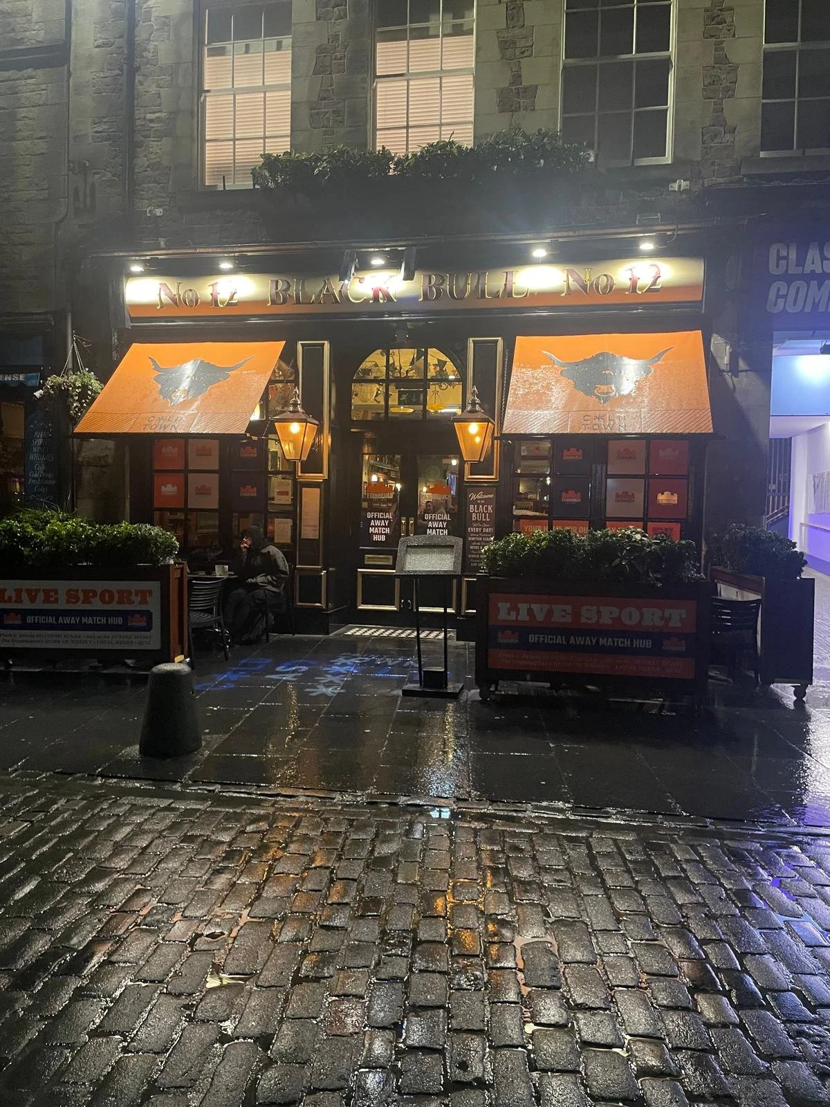 Exterior view of a pub called 'No. 12 Black Bull' at night. The entrance has two orange awnings with a bull's head logo. The door has signs indicating an official away match hub and promotes live sports. There are outdoor seating areas with a few peo