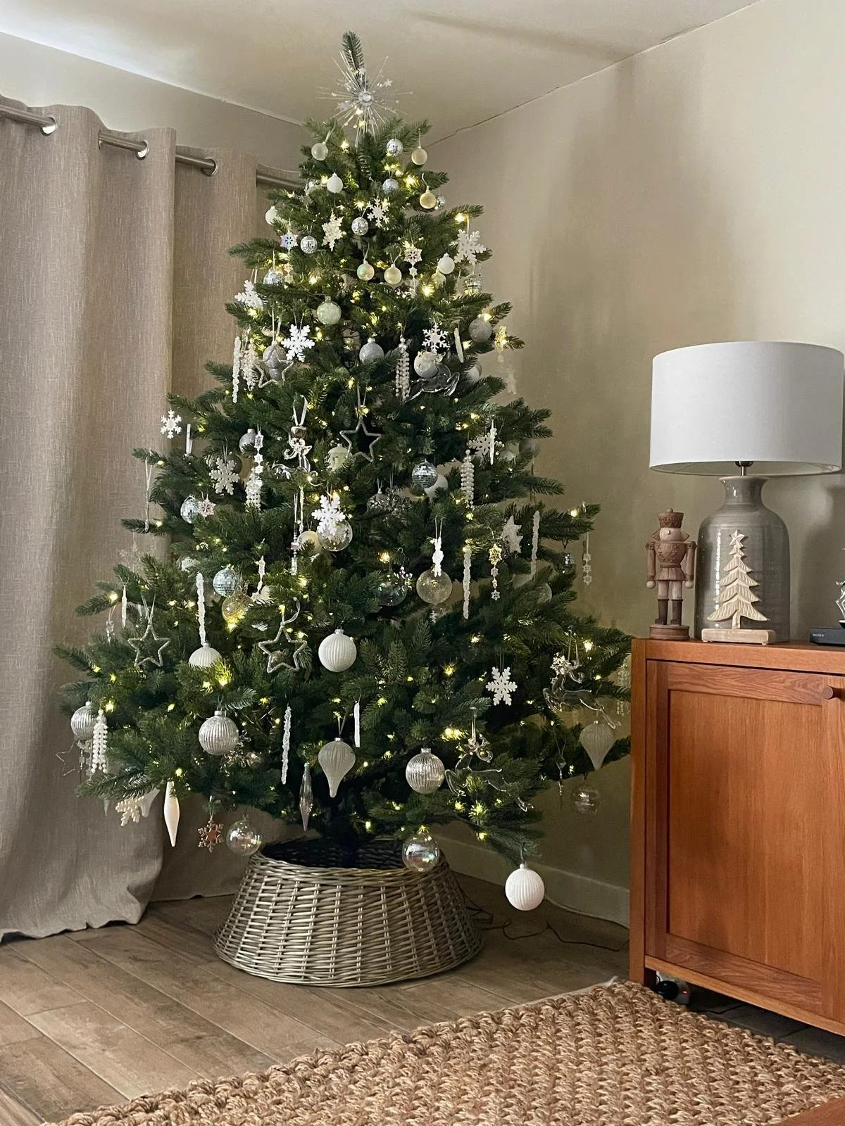Decorated Christmas tree with white and silver ornaments, lights, and snowflake ornaments in a living room corner next to a wooden cabinet with Scandinavian-style decorations, a white lampshade, and a nutcracker figure.