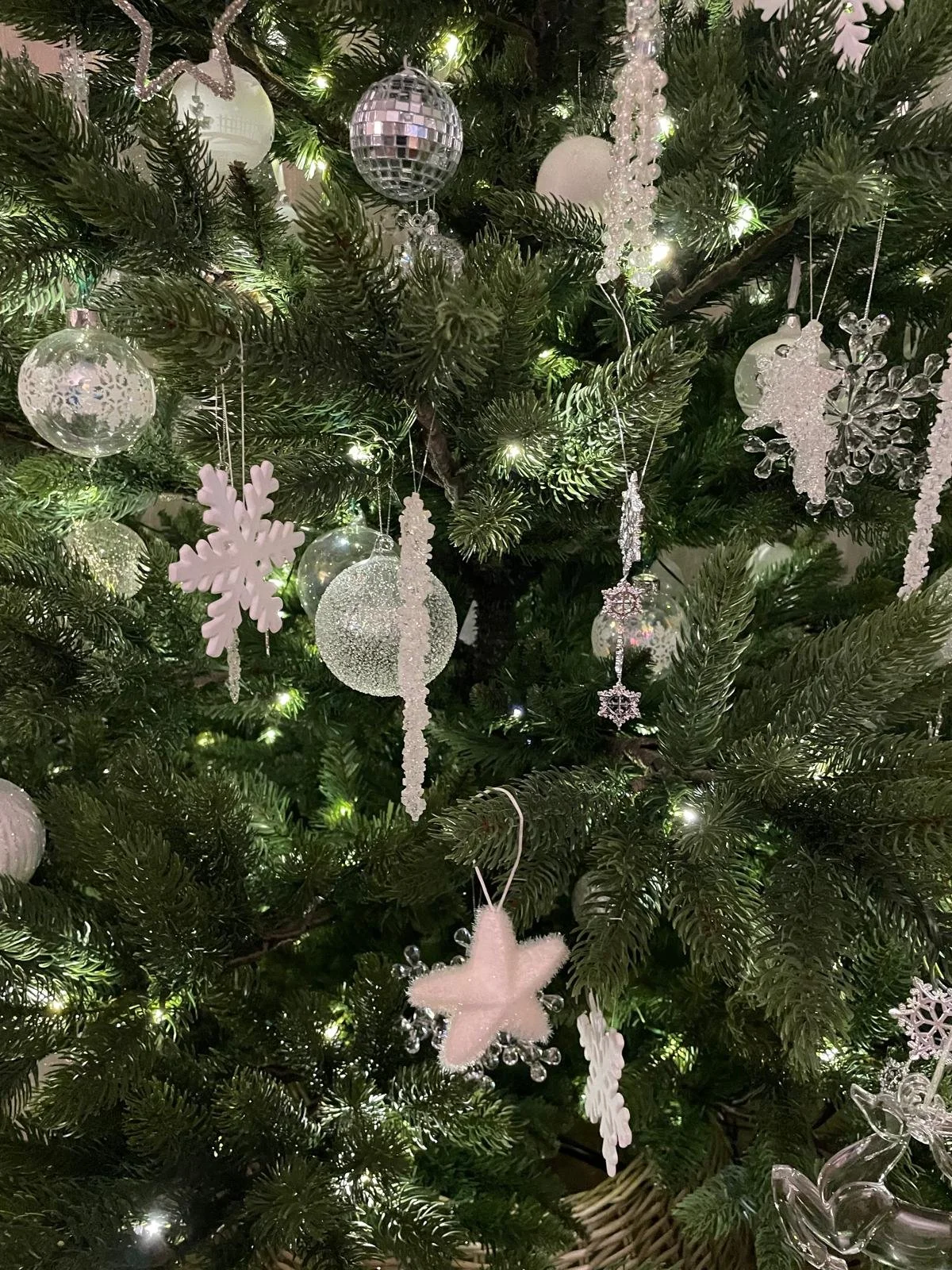 A close-up of a decorated Christmas tree with white and clear ornaments, including snowflakes, balls, stars, and a plush star at the bottom. The tree is illuminated with small white lights.