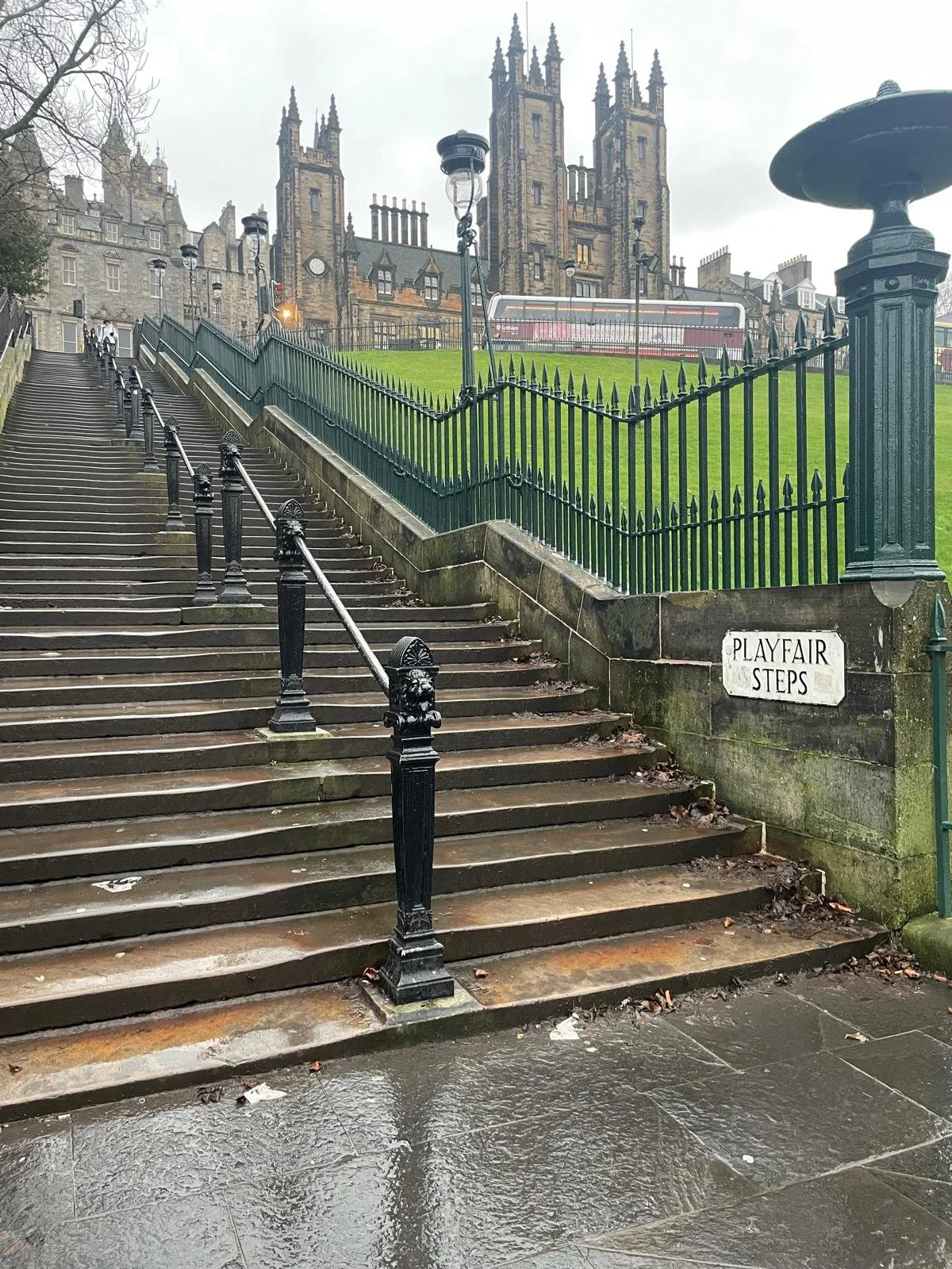 Stone stairs with a black iron railing leading up to a grassy area surrounded by a black iron fence, with a historic castle-like building in the background and a sign reading "Playfair Steps."