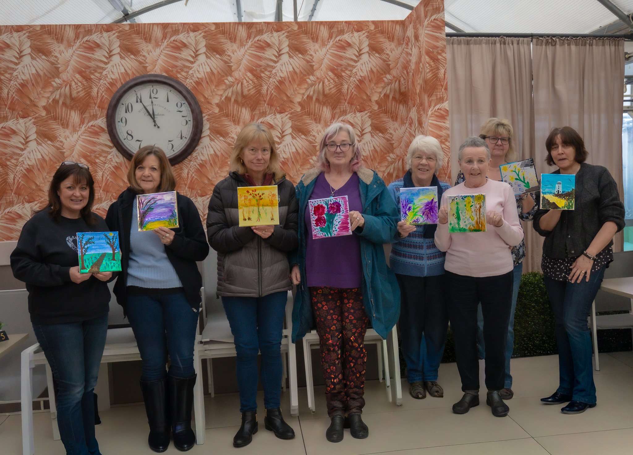 Group of nine women displaying colorful paintings in an indoor setting with a large clock and tropical patterned wallpaper in the background.