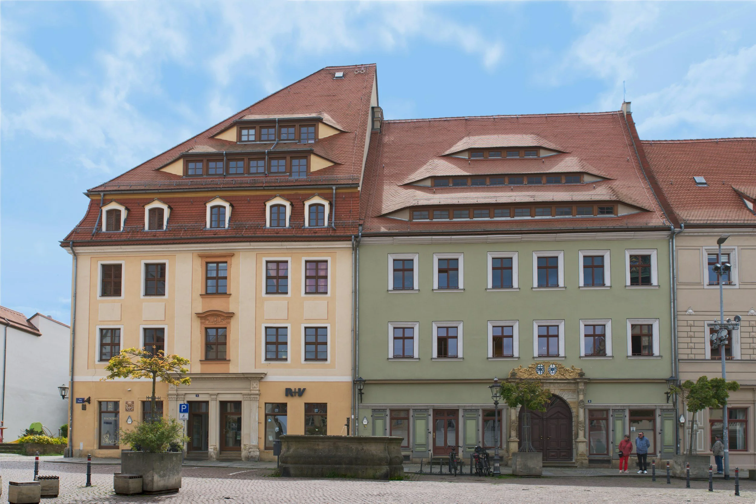 Historische bunte Gebäude mit unterschiedlichen Dächern und Fenstern, eine kleine Parkanlage mit Bäumen im Vordergrund, zwei Personen spazieren, blauer Himmel mit wenigen Wolken.
