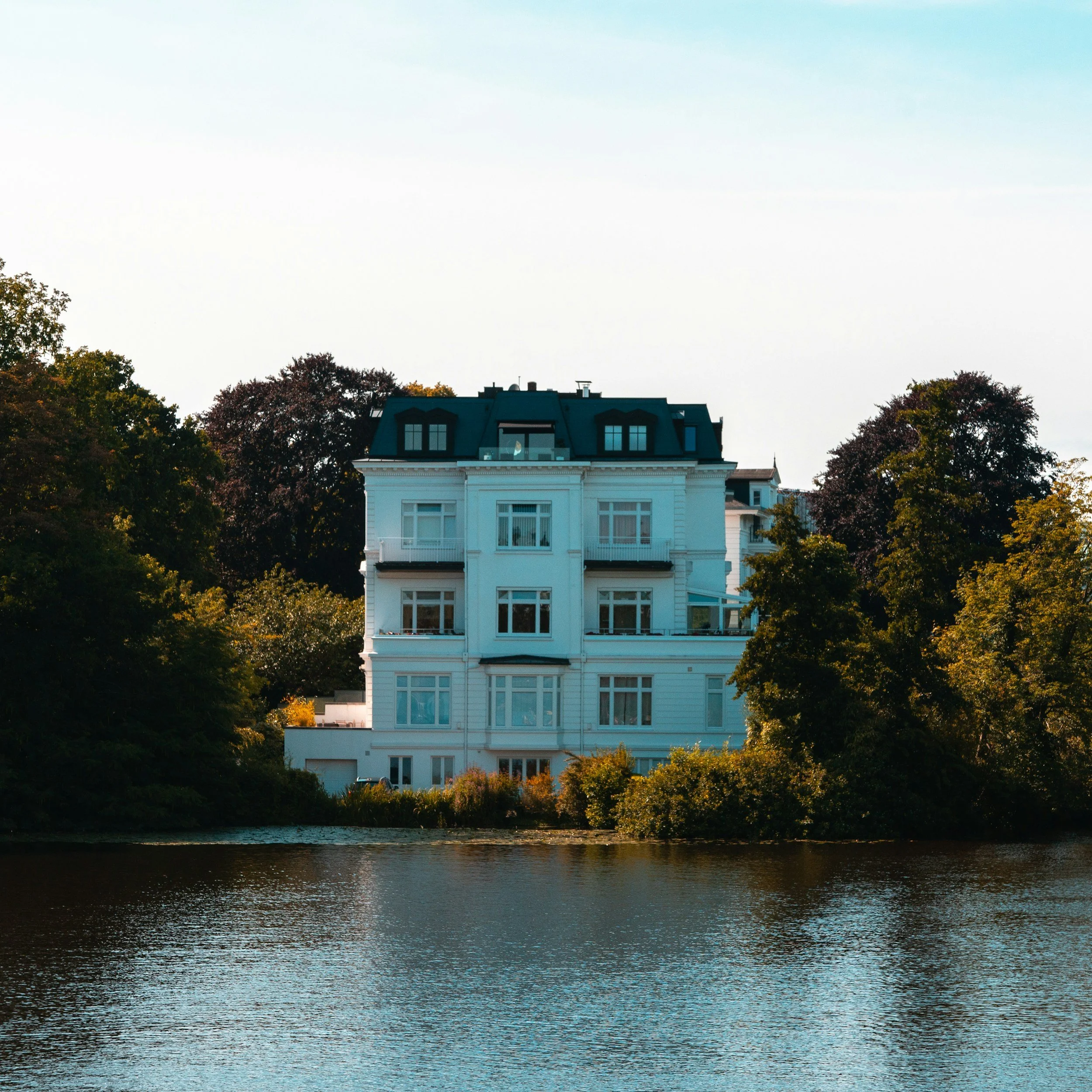 Ein großes weißes Gebäude mit dunklen Dächern und vielen Fenstern, umgeben von Bäumen mit Herbstlaub, und einem Fluss im Vordergrund.